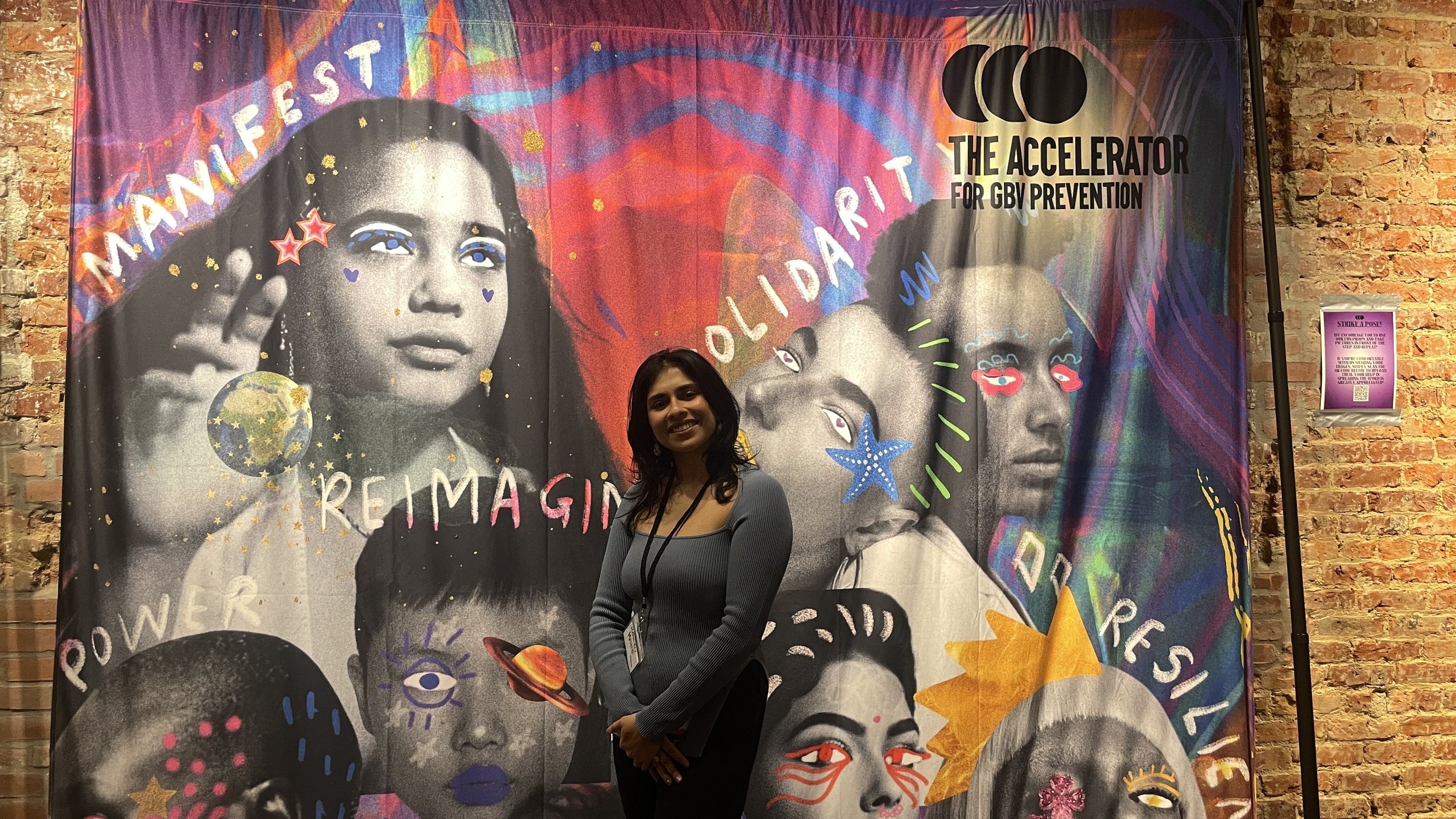 A smiling woman standing in front of a colorful, artistic banner that features black-and-white images of diverse women with decorative doodles and words like 'empower,' 'reimagine,' 'bold,' 'resilience,' and 'manifest.' The banner is titled "The Accelerator for GBV Prevention" and is set against a brick wall.