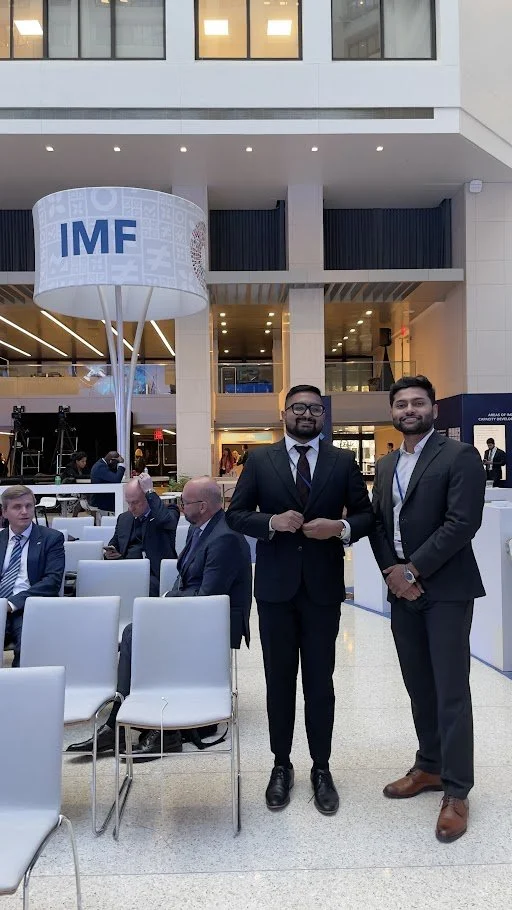 Two men in business suits standing inside a conference center with an IMF sign overhead, surrounded by seated attendees in formal attire.