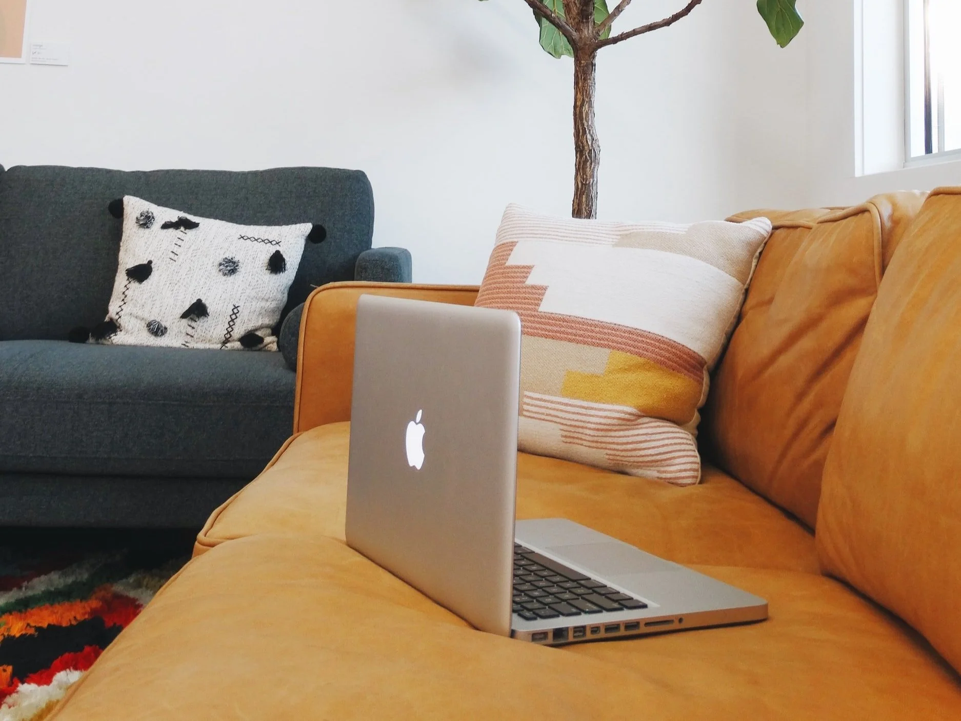 A cozy living room with a mustard-colored leather sofa, a gray fabric armchair, and a colorful woven pillow. A silver MacBook is open on the sofa, and there are large window and a potted plant in the background.