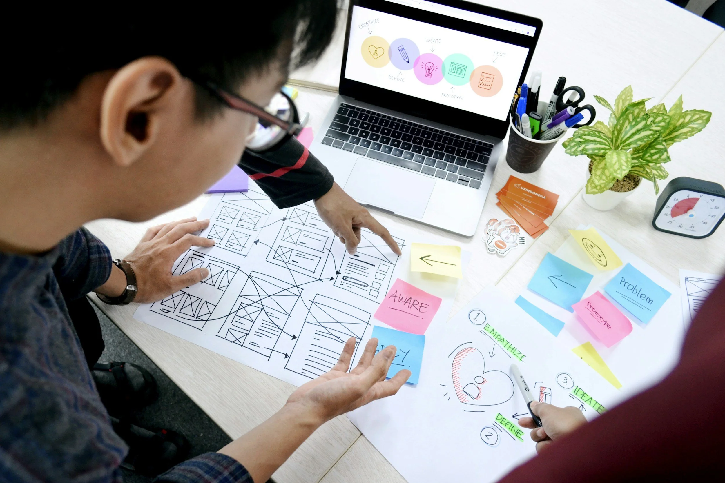 People working on a project with sketches, sticky notes, and a laptop with colorful flowcharts and diagrams, in a collaborative workspace.