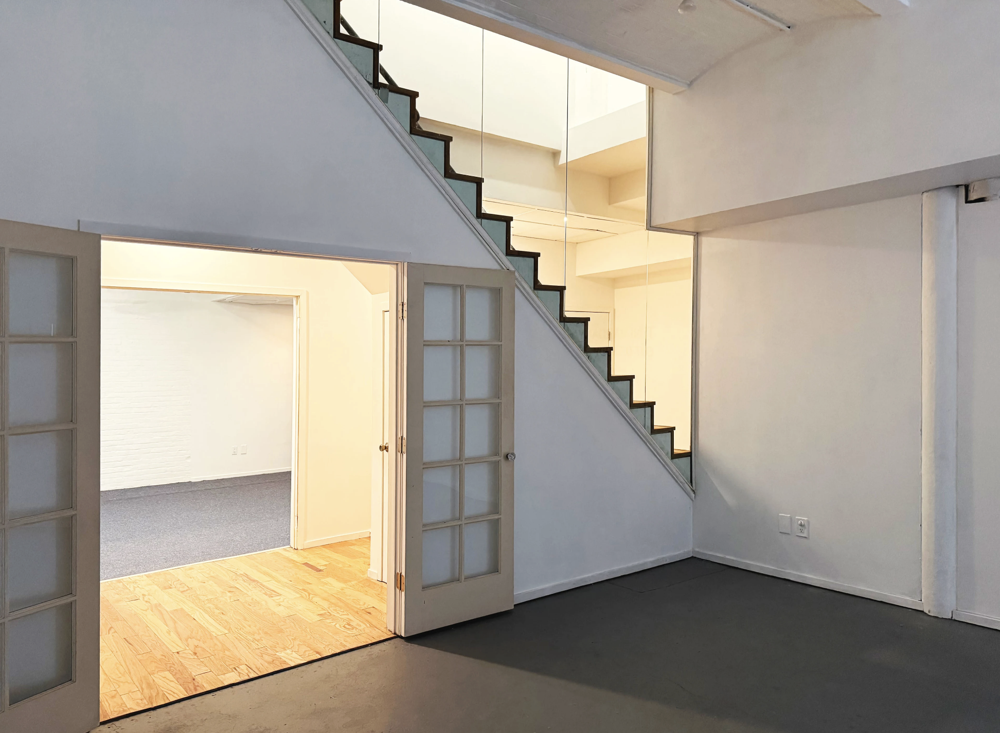 Empty indoor space with a staircase leading upward, white walls, double glass doors opening into a room with a carpeted floor, and a wall with electrical outlets.