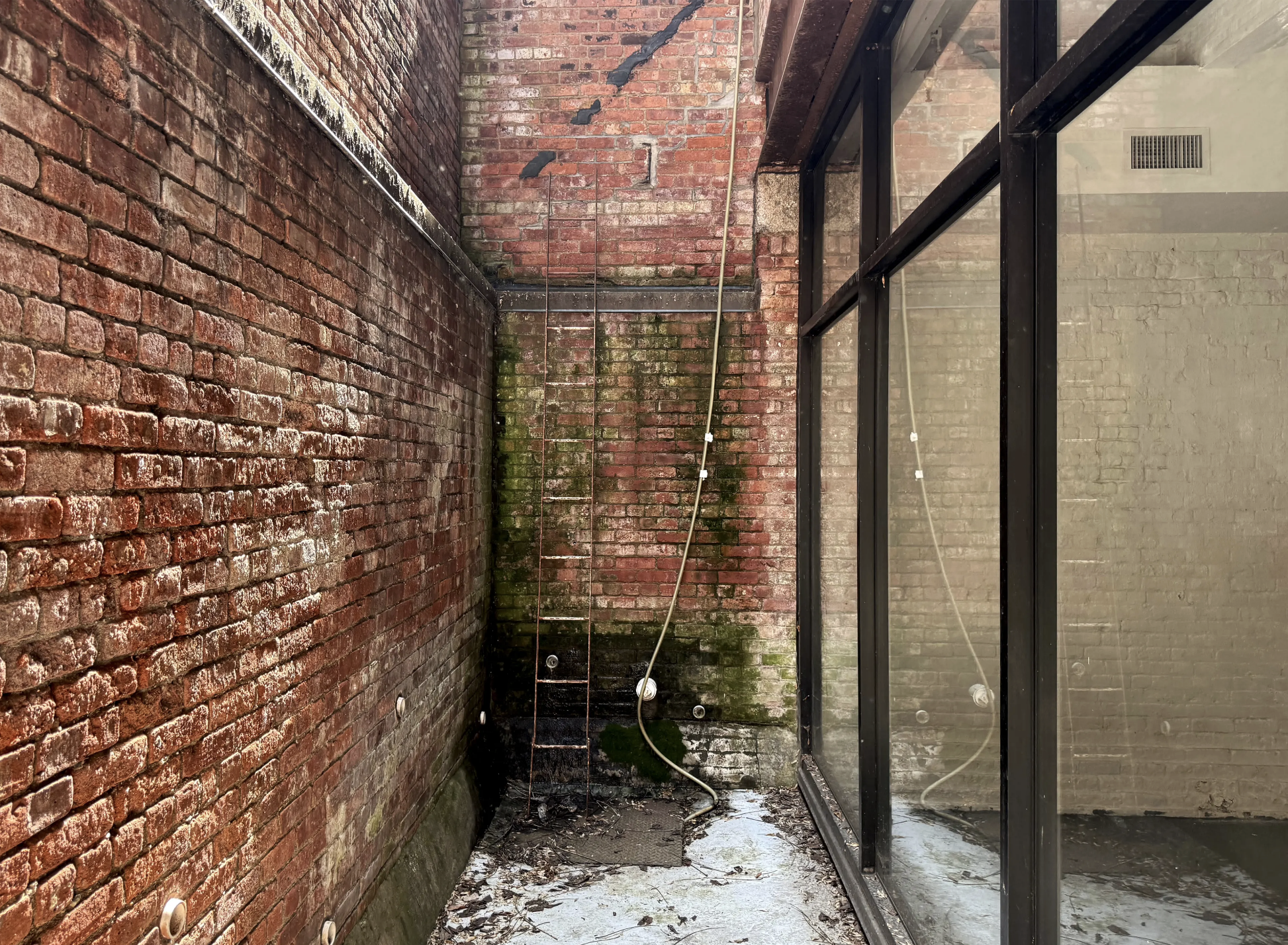 An outdoor corner with old red brick walls, a ladder, and a glass wall reflecting the brick wall.