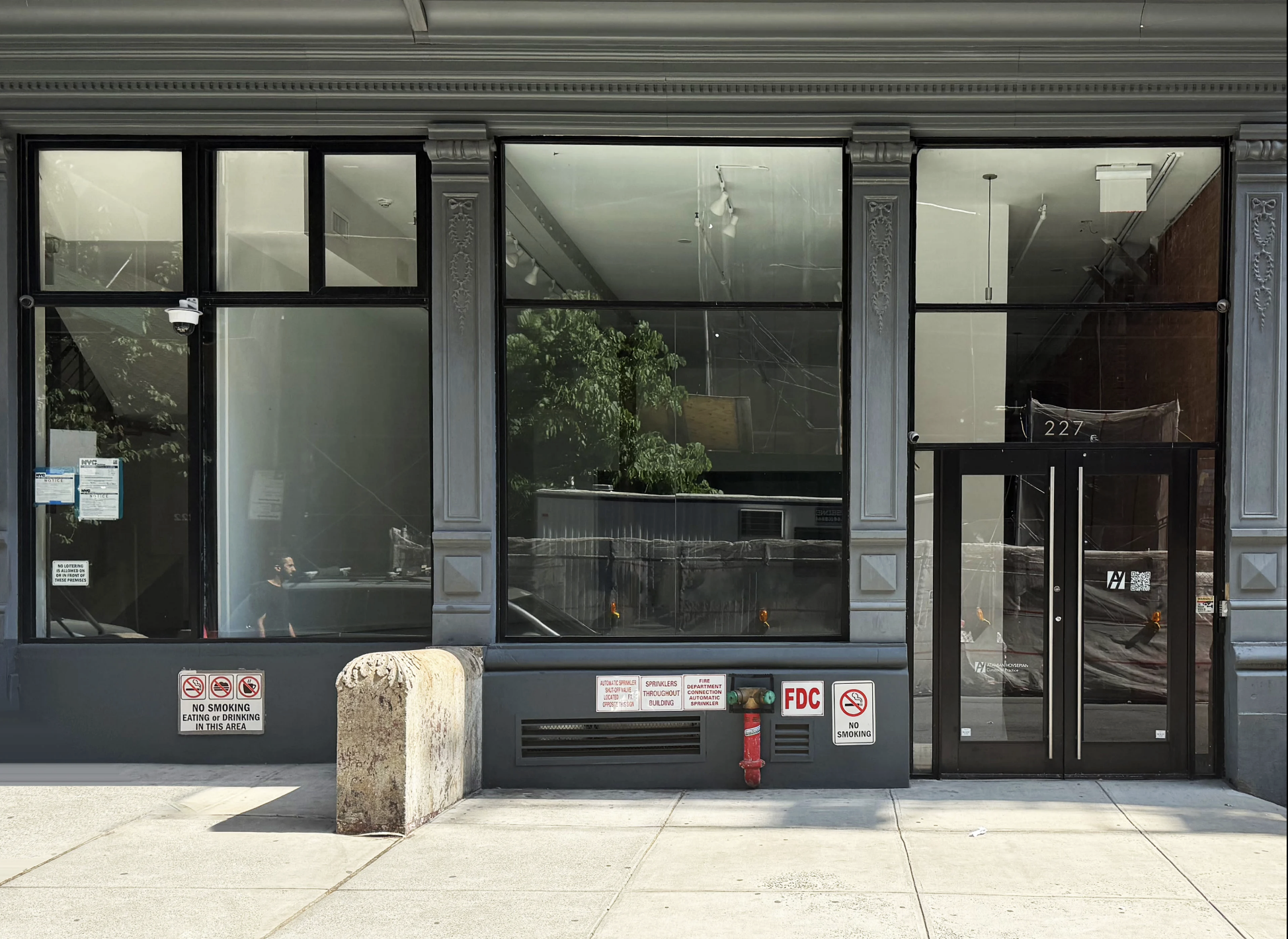 Empty storefront with large glass windows and black frame door, several warning signs, and a fire hydrant outside. Reflection of trees and interior are visible on the glass.