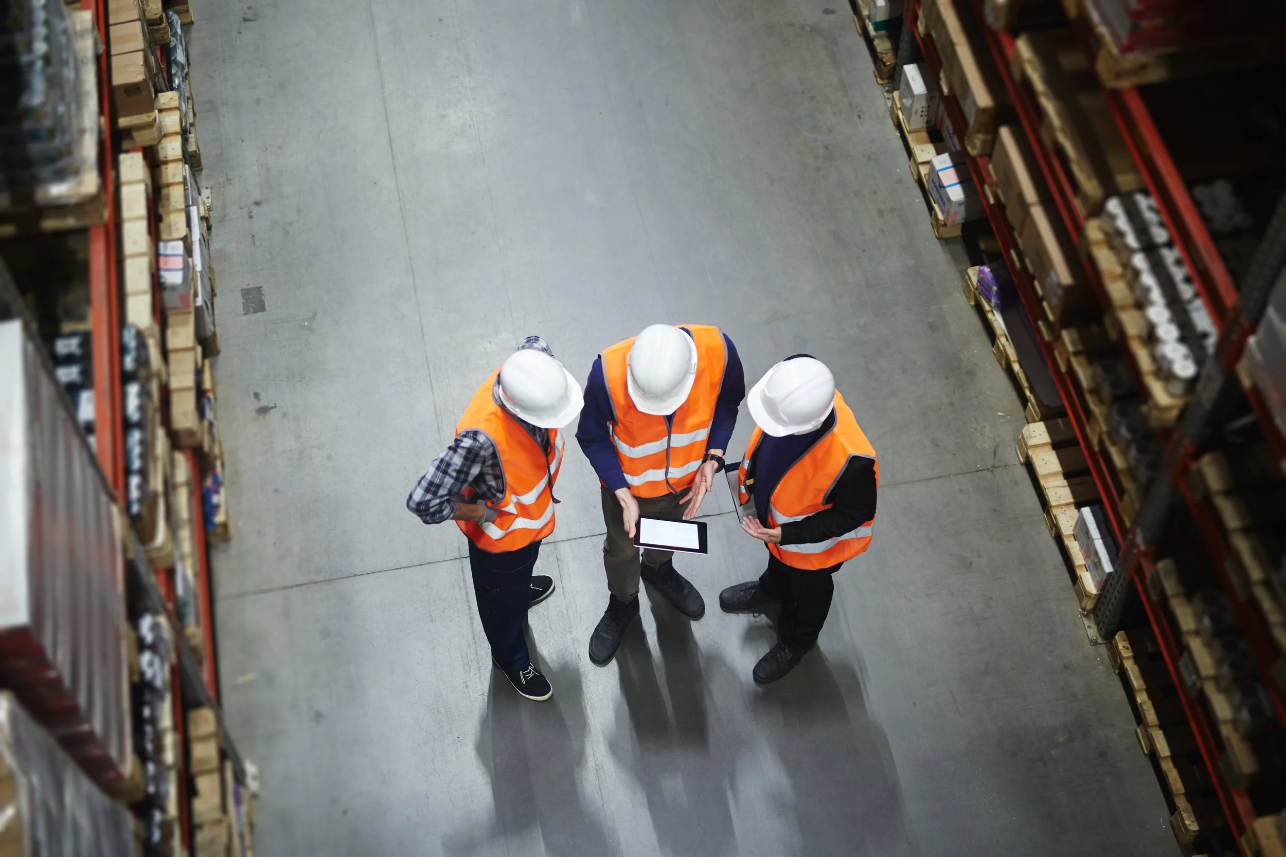 Three workers wearing orange safety vests and white hard hats standing in an aisle of a warehouse, looking at a tablet device.