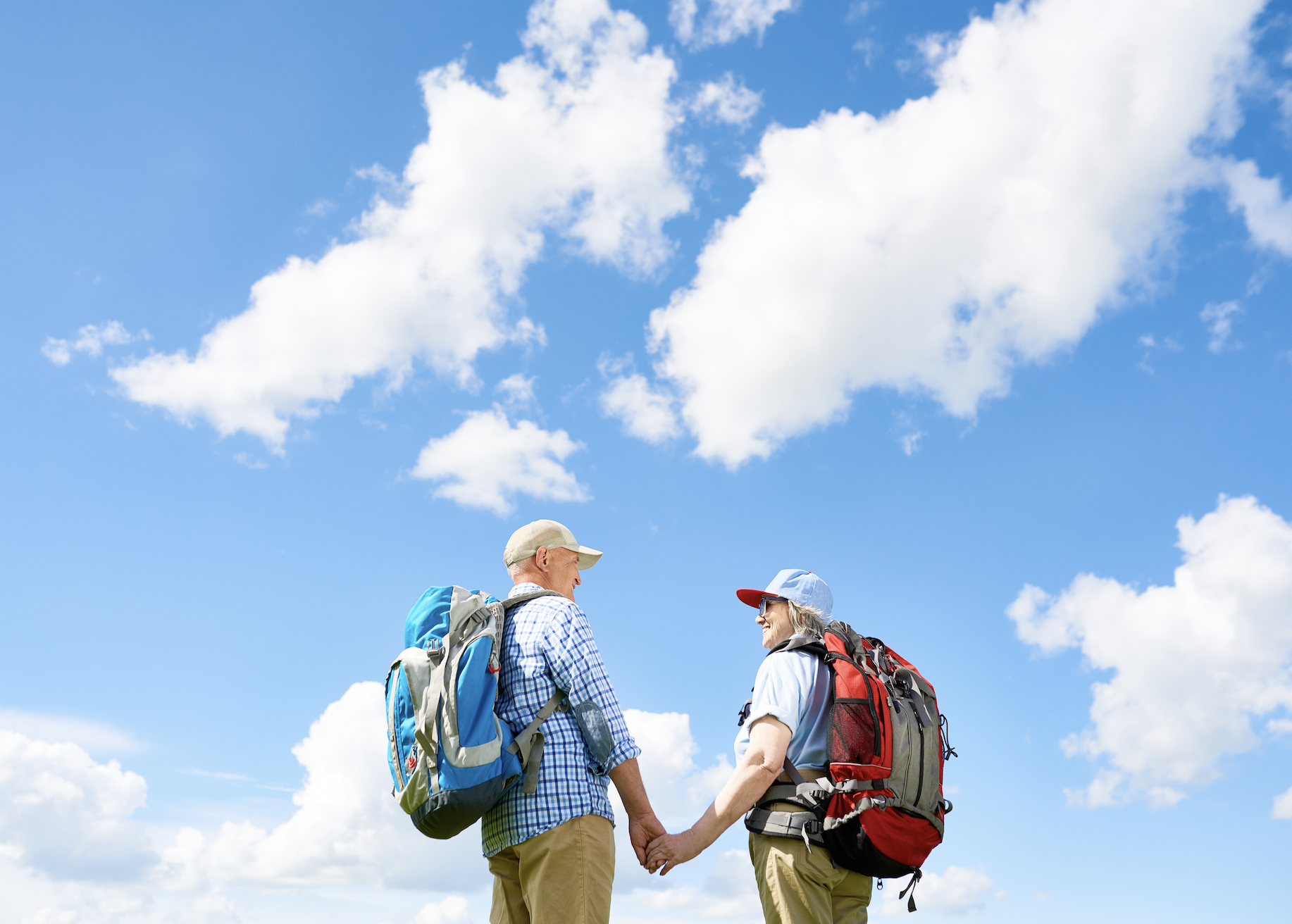 An elderly couple holding hands, standing outdoors with a blue sky and white clouds, both wearing backpacks and casual outdoor clothing.