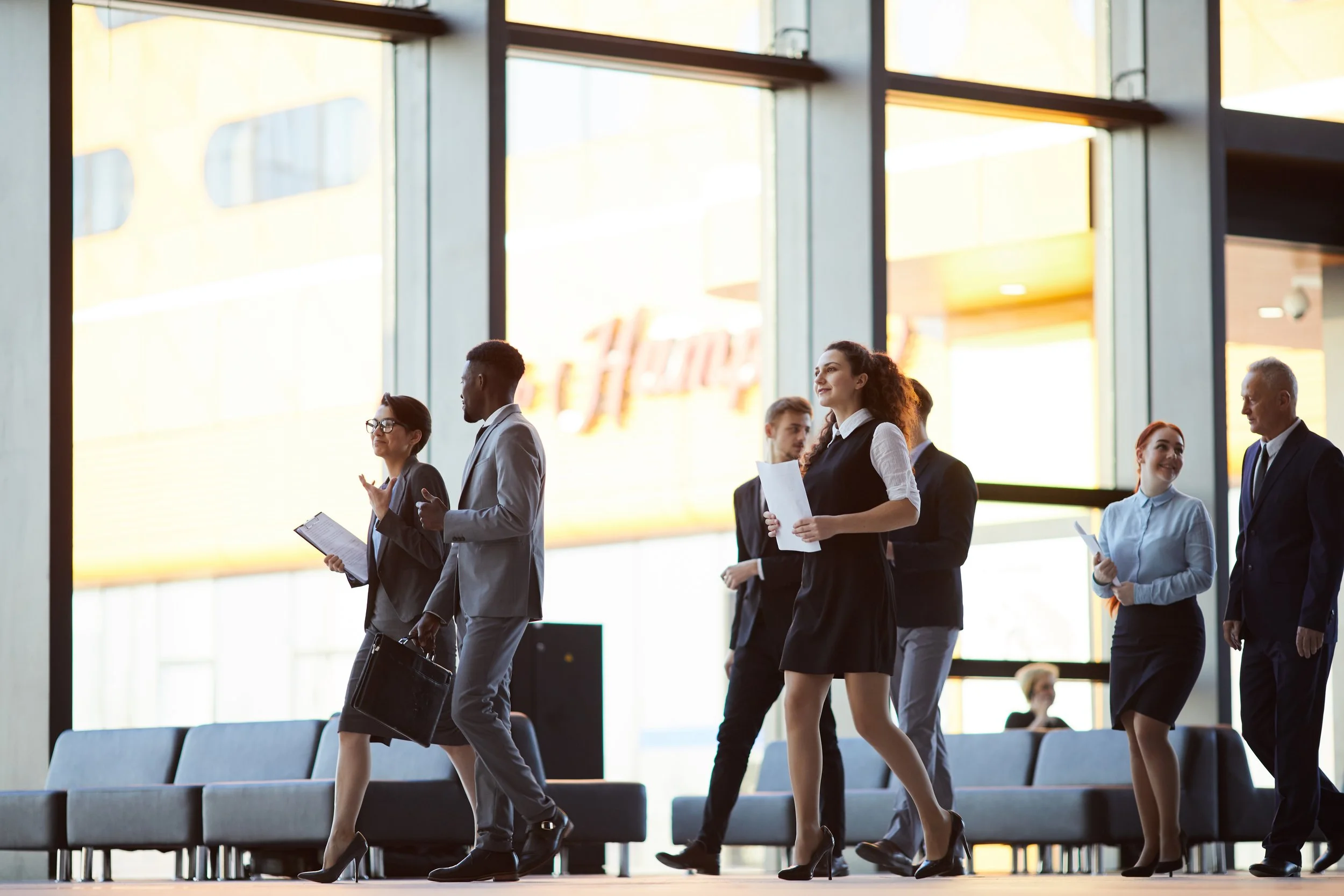 Business professionals walking through a modern airport terminal, some engaged in conversation and holding documents or briefcases.
