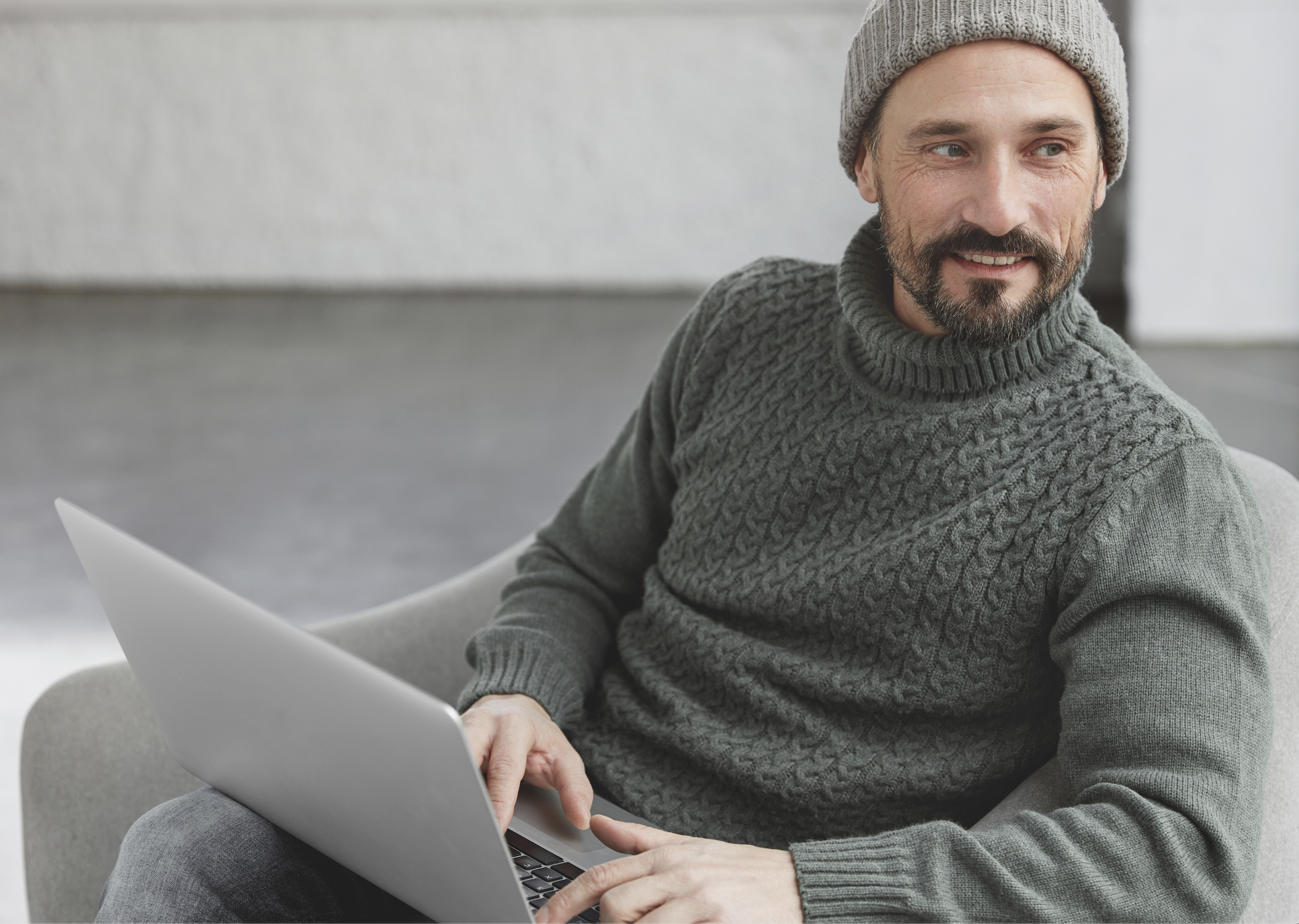 A man wearing a grey knit sweater and a grey beanie sitting on a light-colored couch, using a silver laptop in a modern indoor setting.