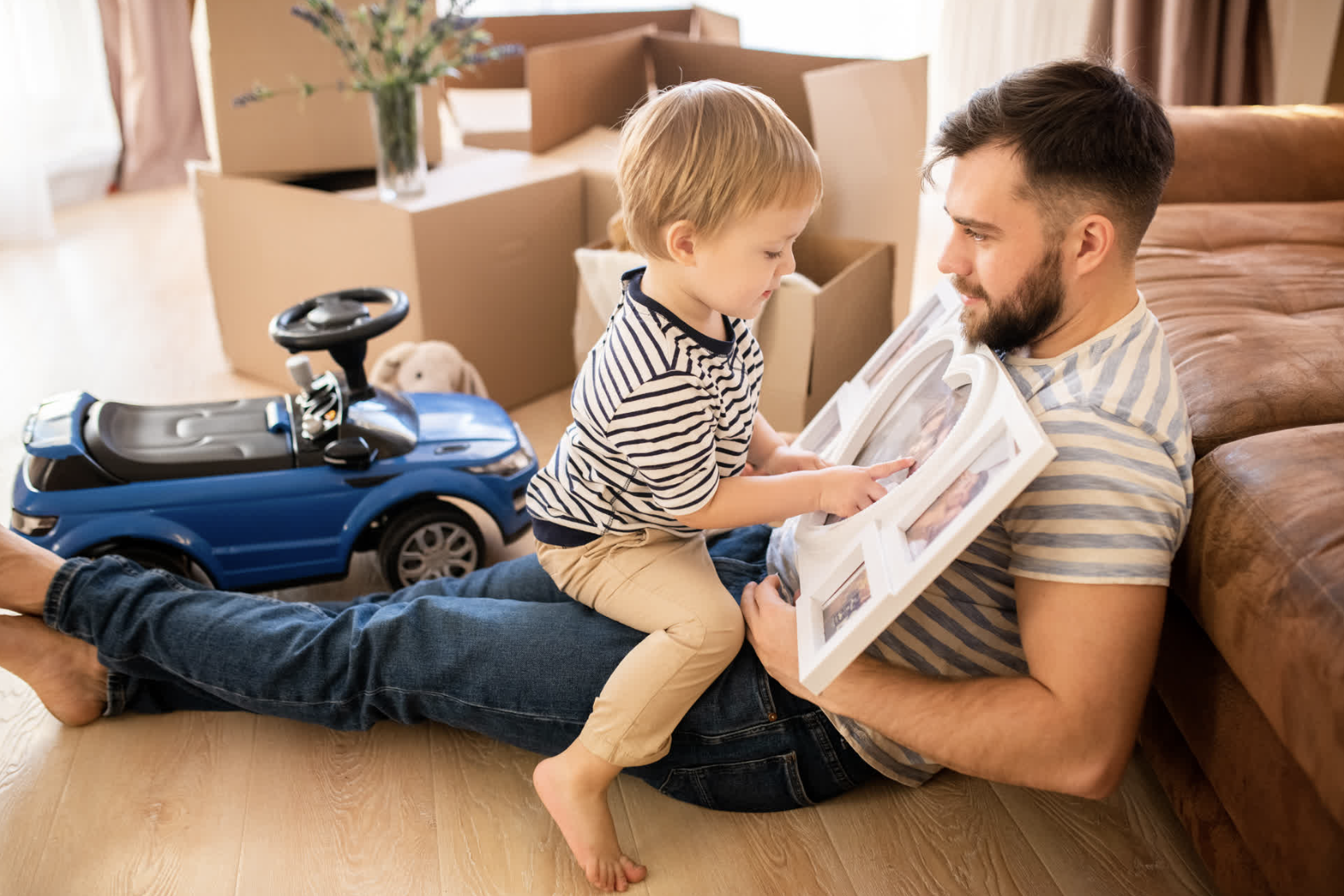 A young boy and a man, likely his father, lying on the floor surrounded by moving boxes, with a toy car and a teddy bear in the background. The boy is pointing at a framed photo or artwork held by the man.