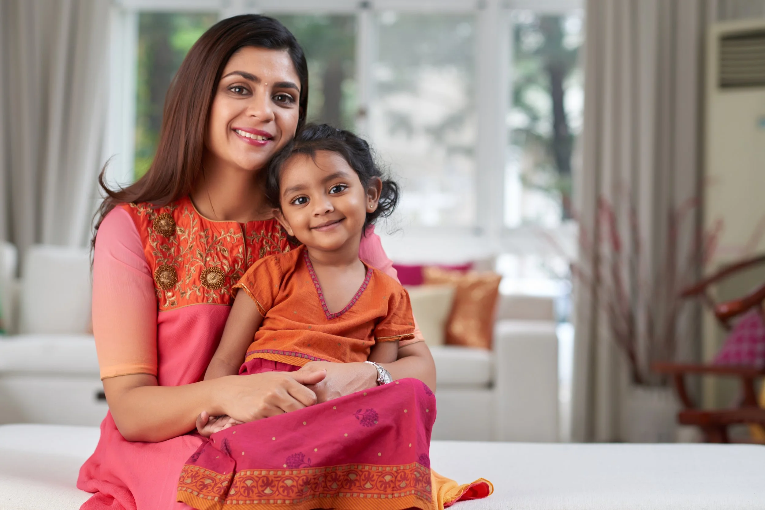 A woman and a young girl sitting on a white sofa in a bright living room, smiling. The woman has brown hair and is wearing a pink and orange traditional dress with embroidery. The girl has dark hair, is wearing an orange top and a pink skirt, and is smiling at the camera.