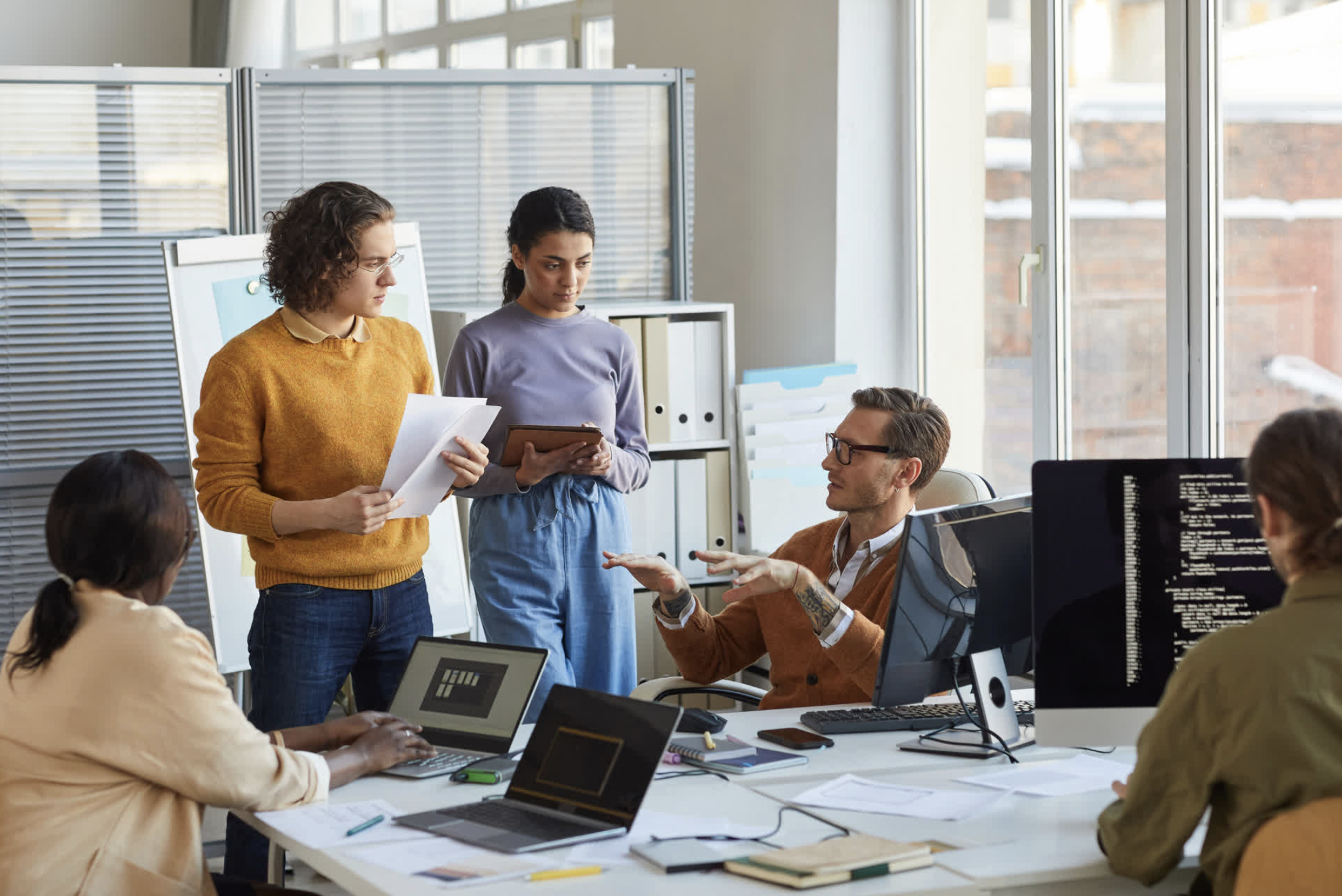 Group of diverse people having a discussion in a modern office with large windows, laptops, and documents on the table.