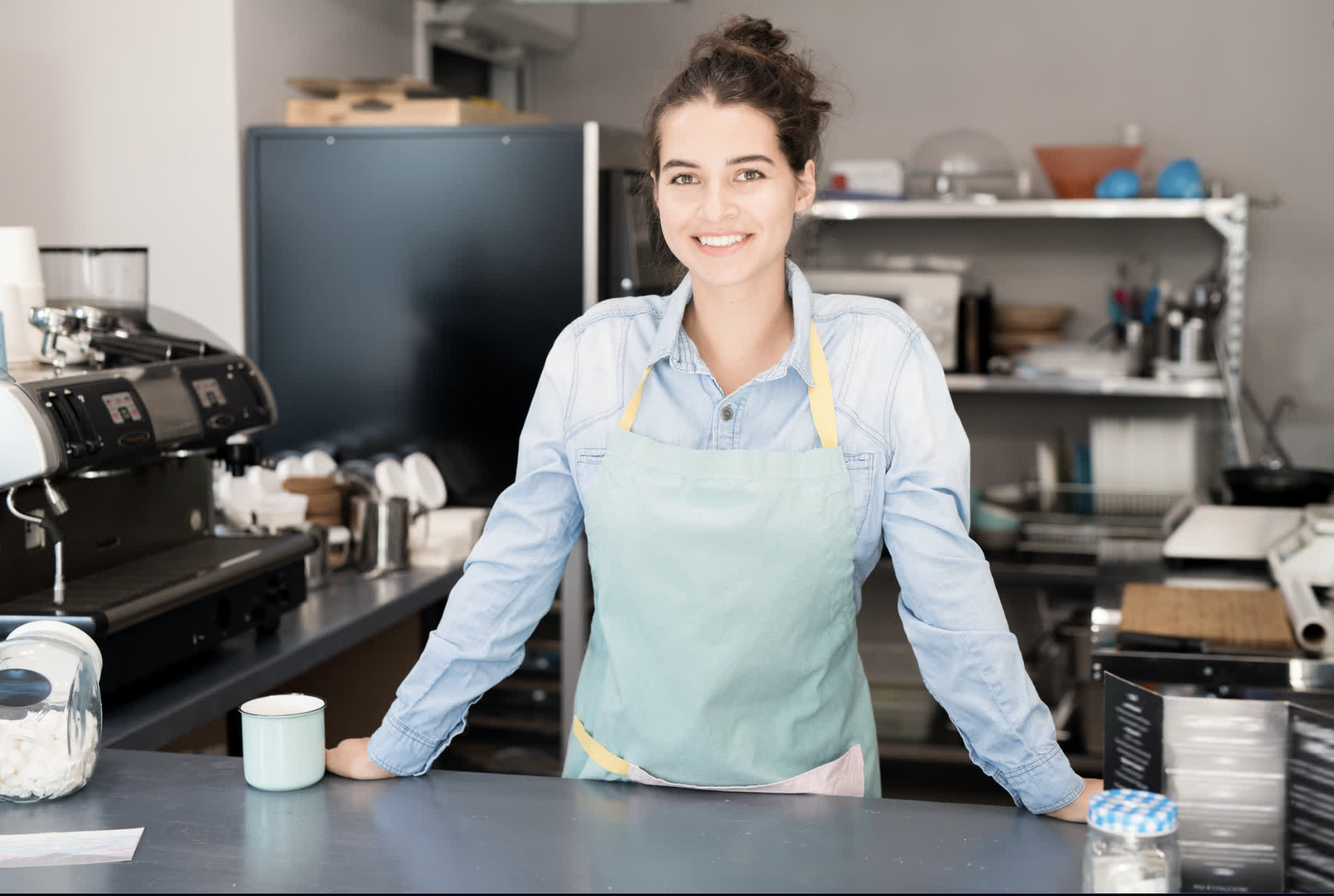 A young woman in a light blue shirt and pastel apron smiling at the camera in a kitchen or bakery setting, with baking supplies around her.