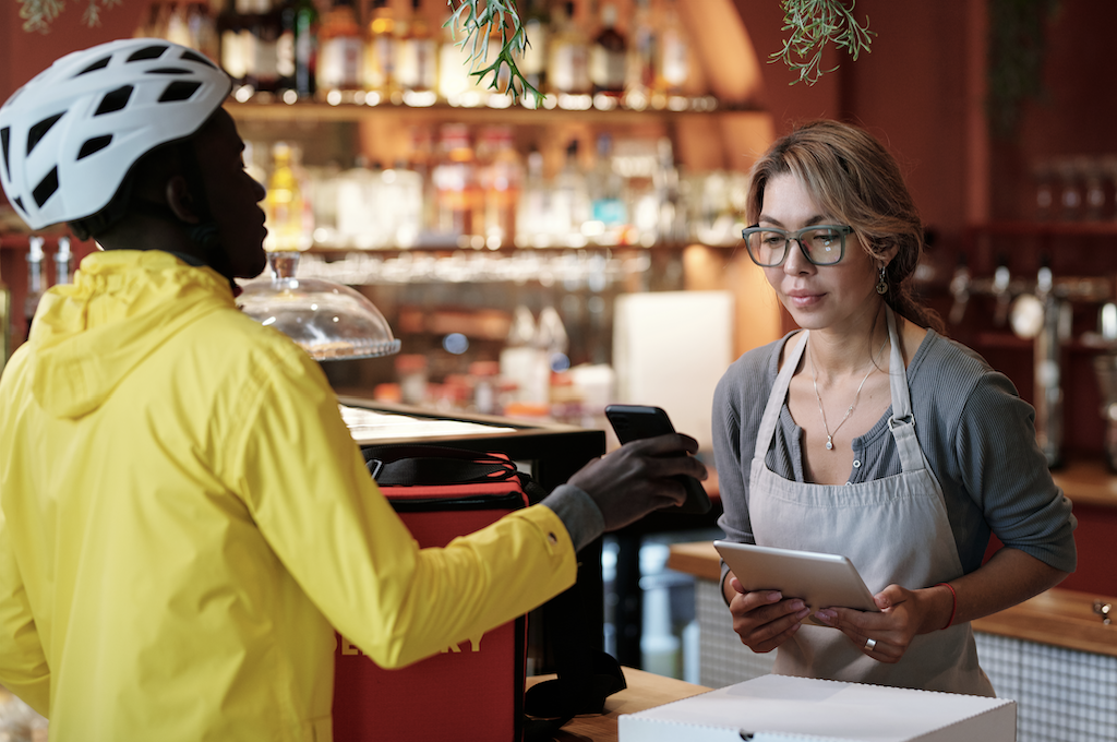 A delivery person in a yellow jacket and helmet handing over a package to a woman at a cafe counter. The woman has glasses, a gray shirt, an apron, and is holding a tablet. There are shelves with bottles in the background.