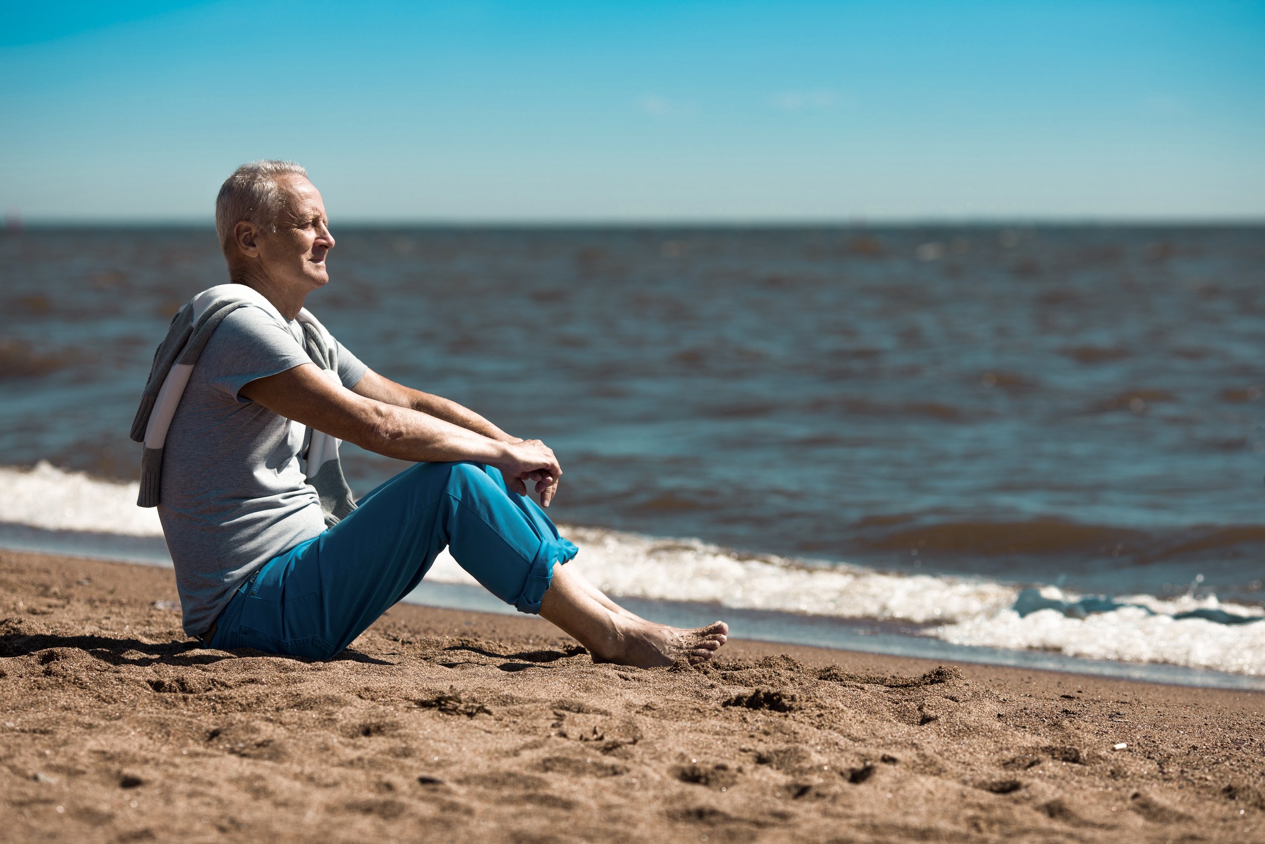 An elderly man sitting on the sandy beach near the ocean, gazing at the water with a relaxed expression.