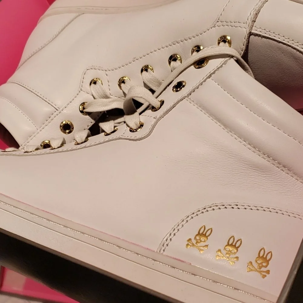Close-up of a white sneaker with gold eyelets and bunny logo detail on the toe box.