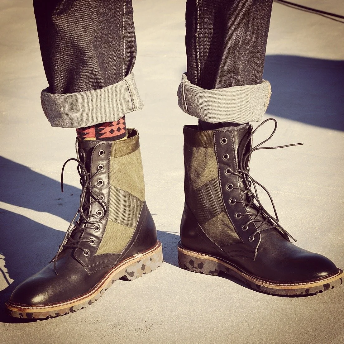 Person's feet wearing hiking boots with patches of olive green fabric, rolled-up jeans, and patterned socks on a sunny surface.