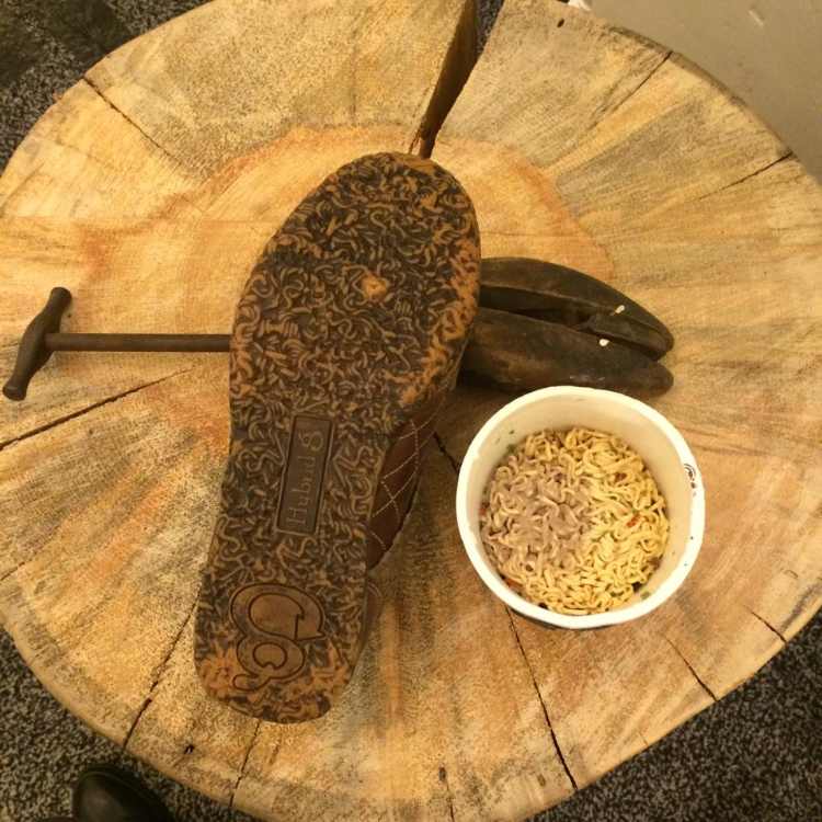A wooden table with a bag of shoes, a bowl of instant ramen noodles, and two bananas.