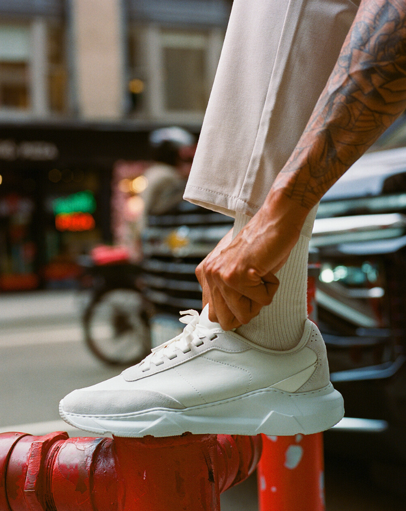 A person adjusting a white sneaker on a red fire hydrant on a city street.