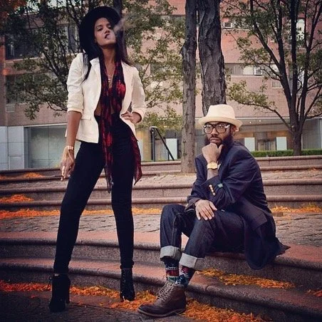 A stylish woman standing with one hand in her pocket and a man sitting on steps outdoors. The woman wears a white blazer, dark hat, and dark pants. The man wears glasses, a light-colored hat, a dark jacket, and jeans with rolled-up cuffs.
