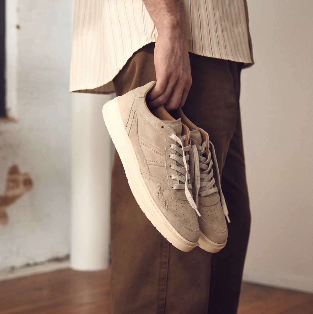 Person holding beige sneakers in their hand, standing indoors with a wooden floor and white wall with exposed brick.