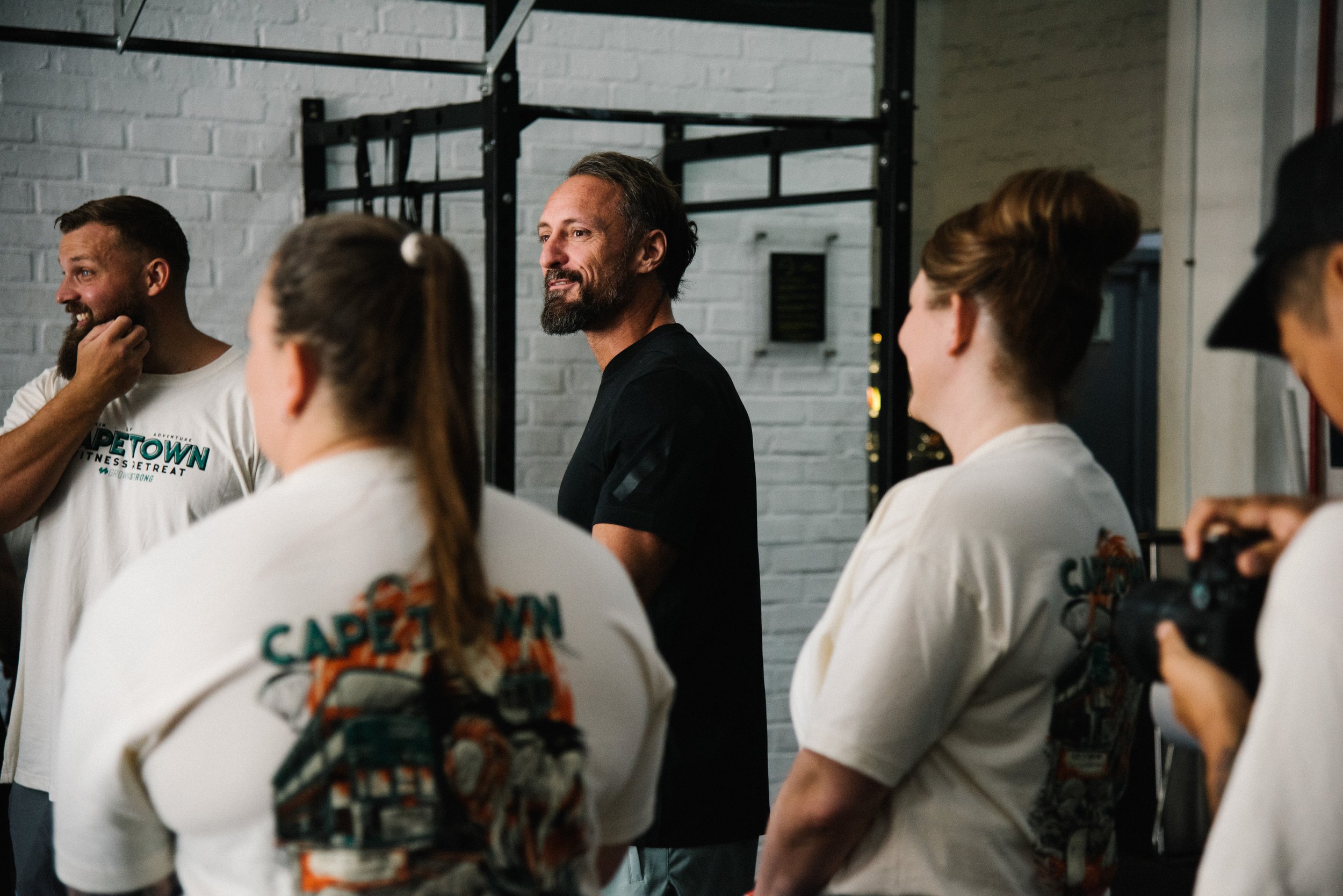 A group of people attending a fitness class indoors, with a man in the center smiling and looking away, and women around him with their backs facing the camera, wearing white shirts with text on the back, in a gym with a white brick wall.