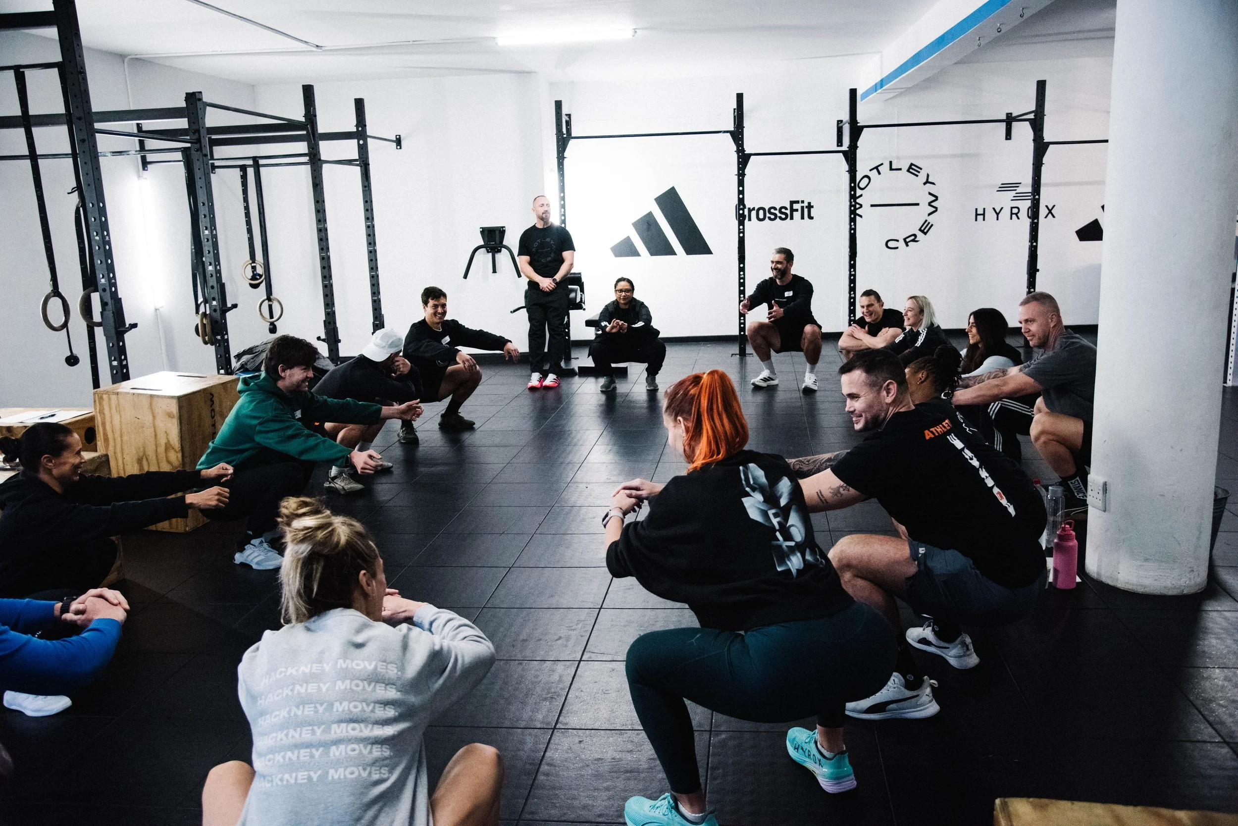 Group of people sitting on the floor in a gym, engaging in a fitness class or discussion, with gym equipment and CrossFit logos visible on the wall.