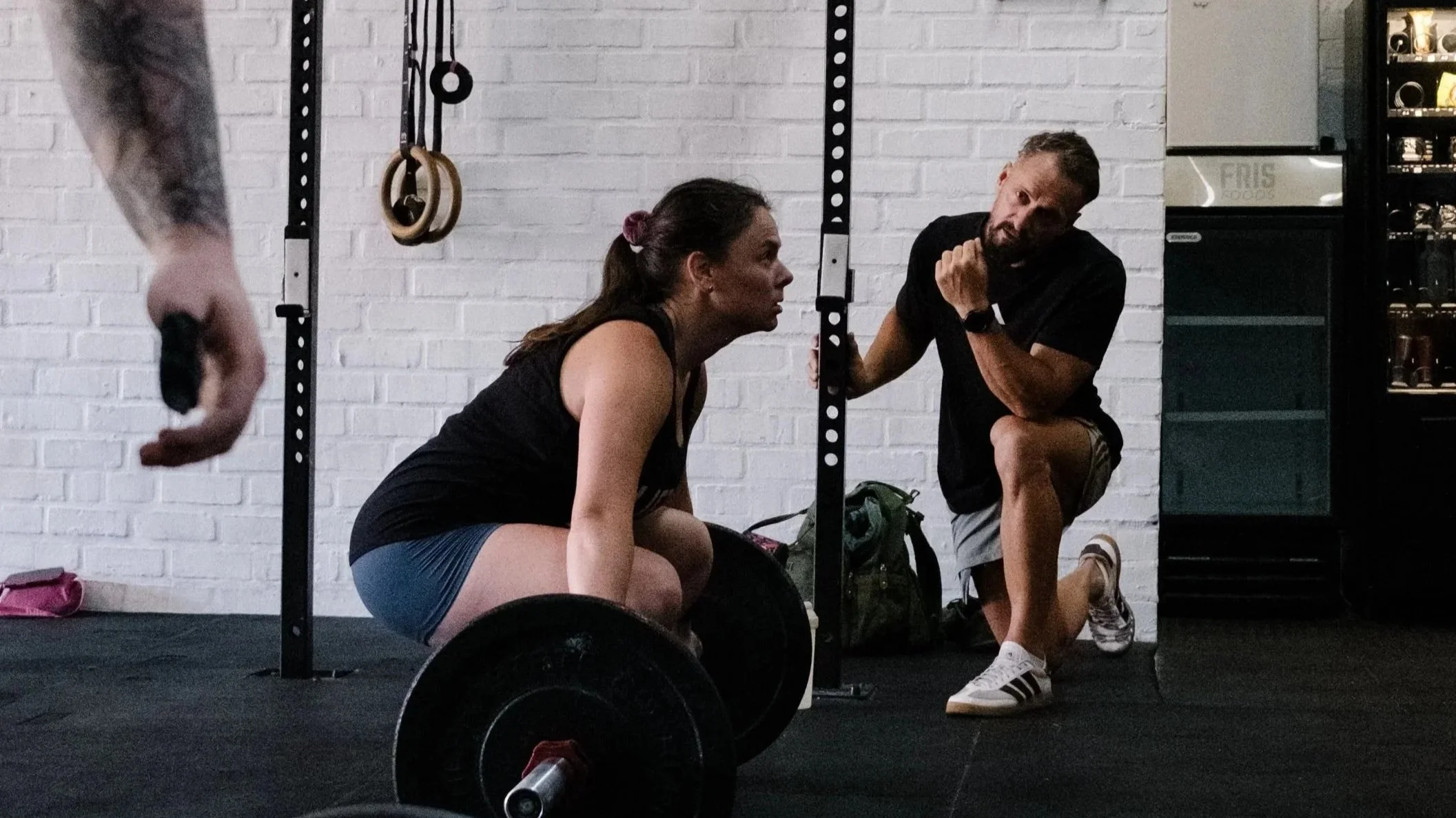 A woman and a man are in a gym, with the woman in a squat position and the man coaching or instructing her, surrounded by gym equipment like rings, weights, and a white brick wall.