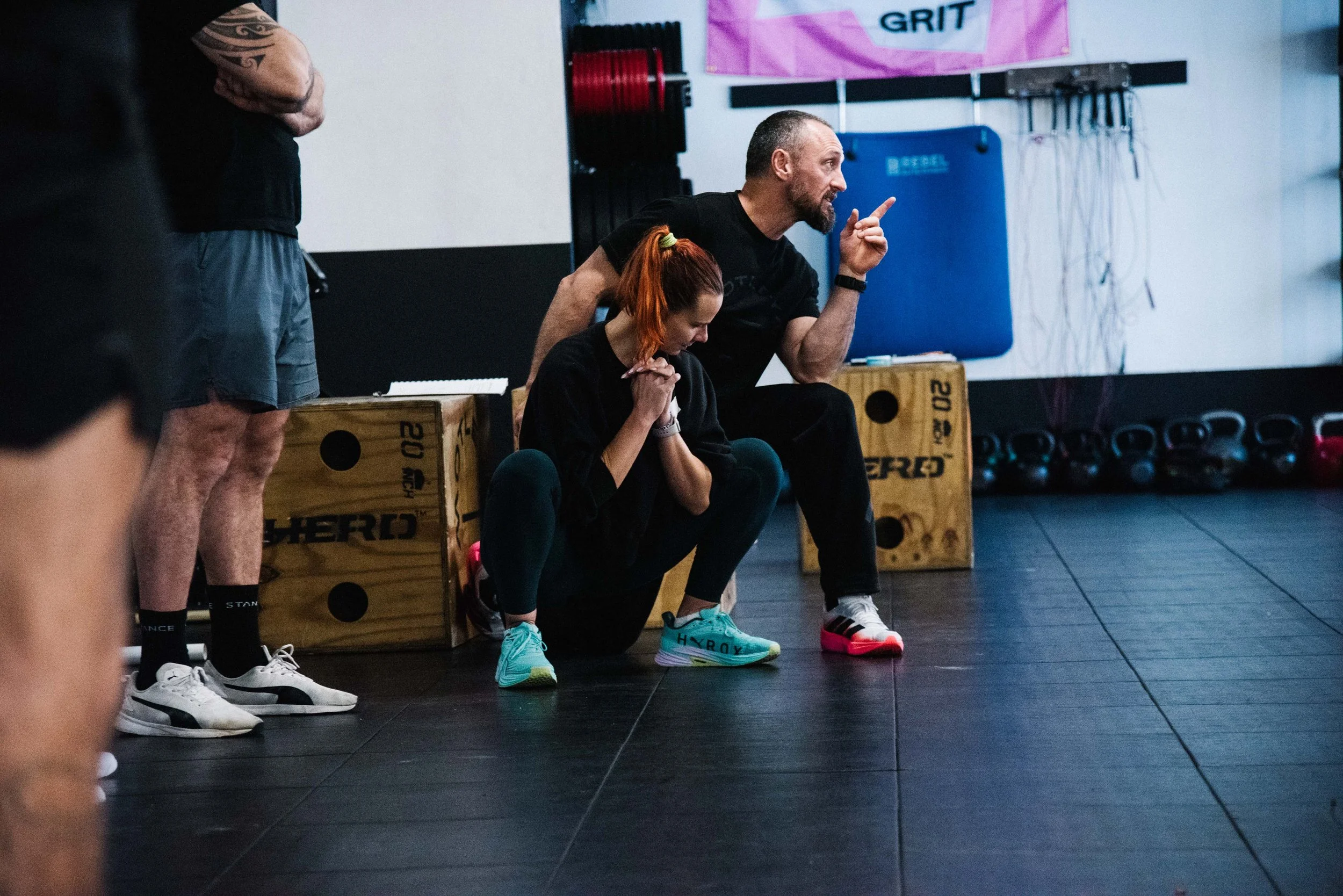 A man and woman in a gym, with the man coaching or instructing the woman during a workout. The woman is crouched down, reaching for her ankles, wearing colorful athletic shoes. Gym equipment and weights are visible in the background.