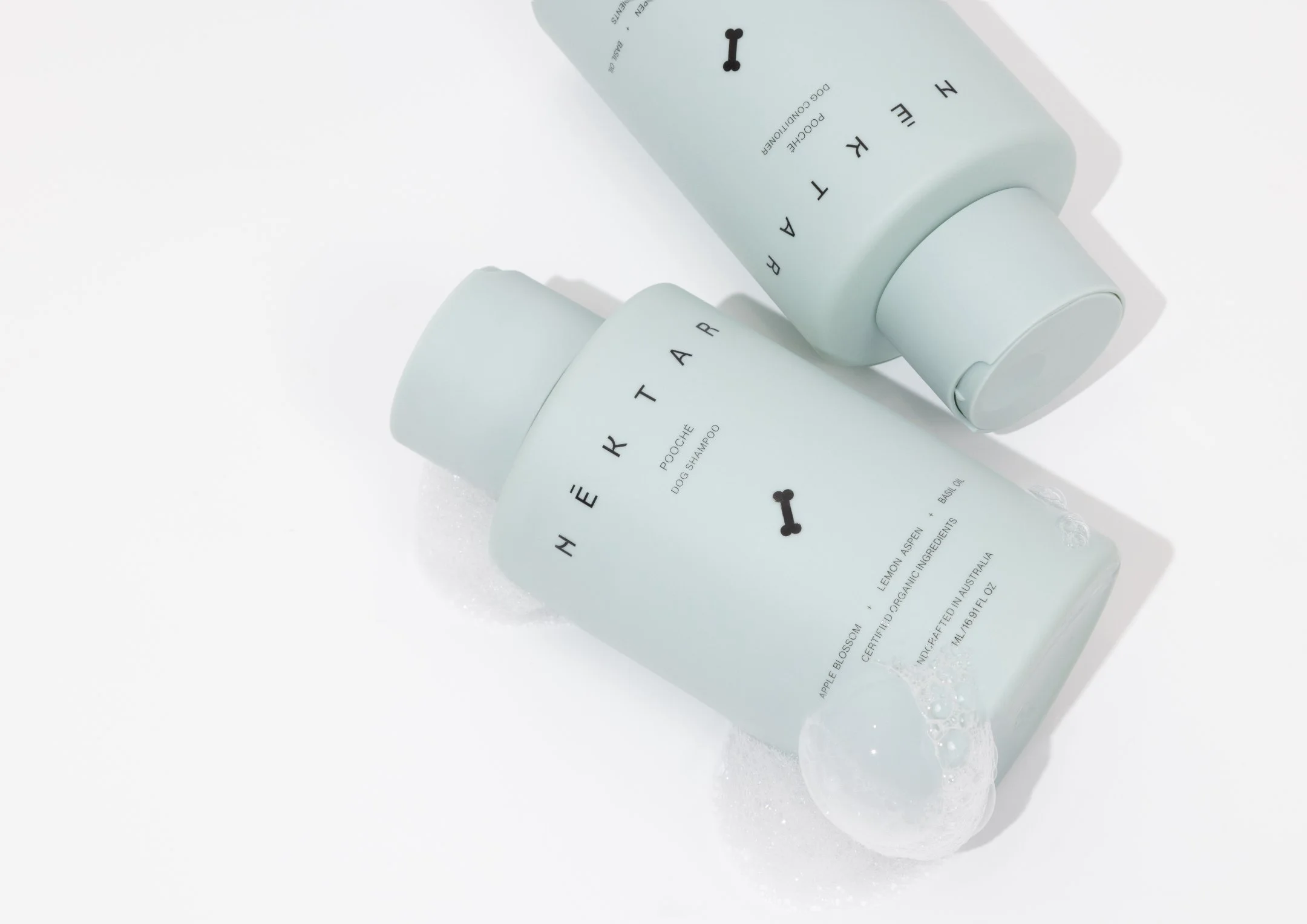 Two pale mint green bottles of Hydrohearth pet shampoo and conditioner, tilted on a white surface with bubbles.