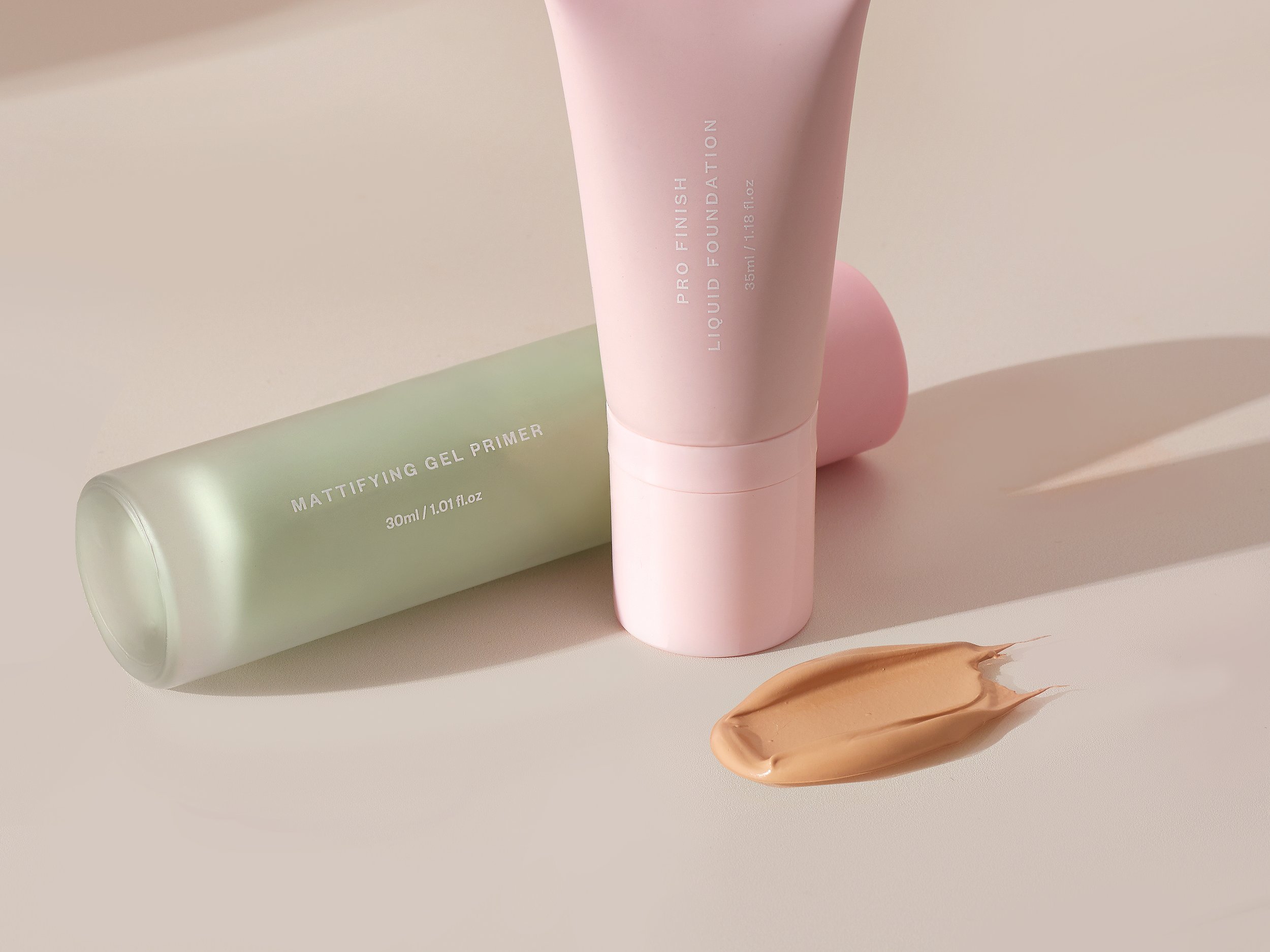 Two cosmetic tubes, one green labeled 'Matteifying Gel Primer' and one pink labeled 'Pro Finish Liquid Foundation,' with a smear of beige foundation on a white surface.