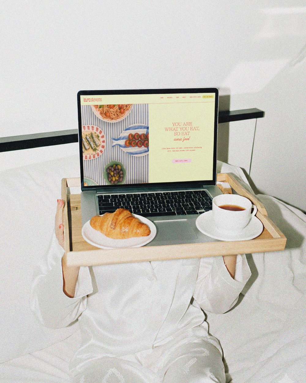 A tray with a laptop, a croissant on a plate, and a cup of tea, held by a person wearing a white robe, in a minimalistic room with a white bed and wall. The laptop screen displays a food website.