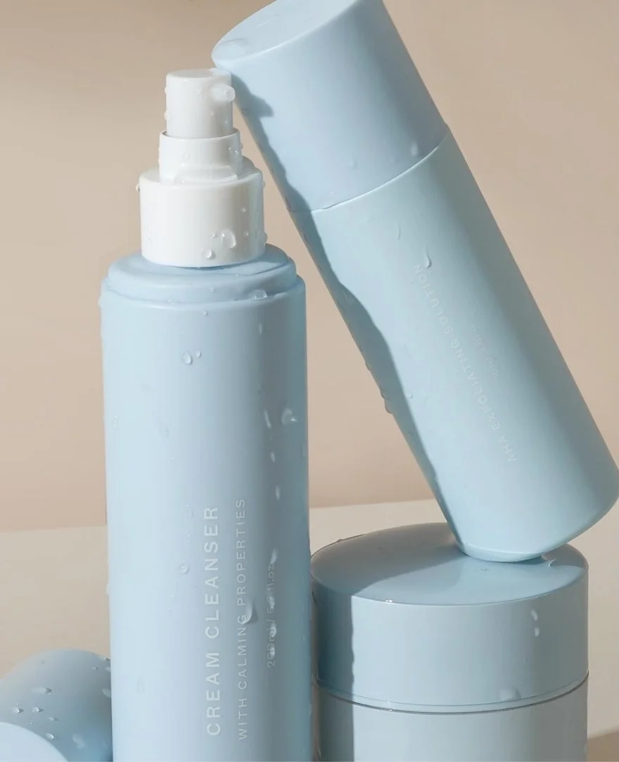 Three light blue skincare bottles, with one open and showing a spray nozzle, are positioned against a neutral background.