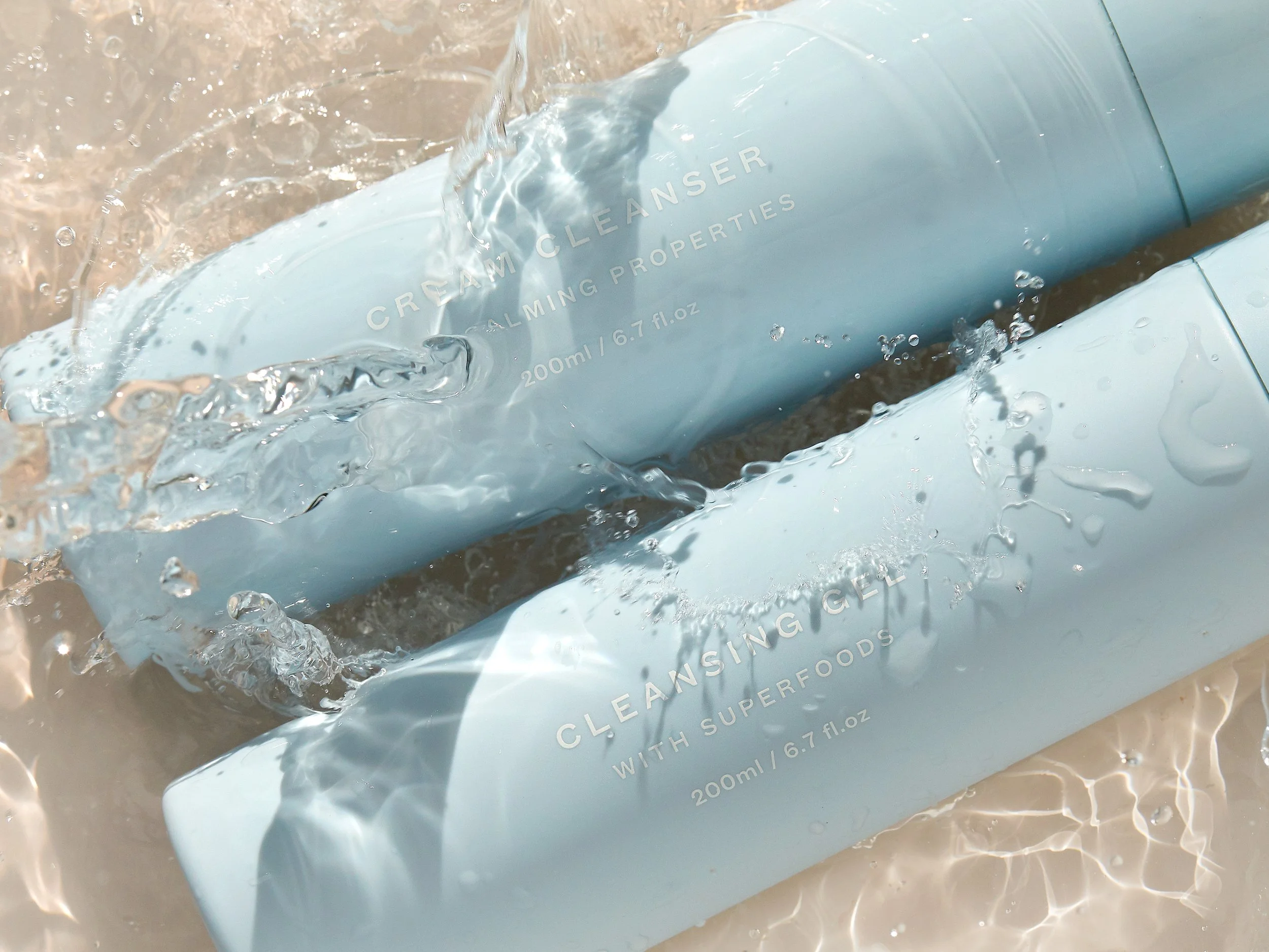 Two light blue bottles labeled 'Cream Cleanser' and 'Cleansing Gel' with water splashing around them on a sandy surface.
