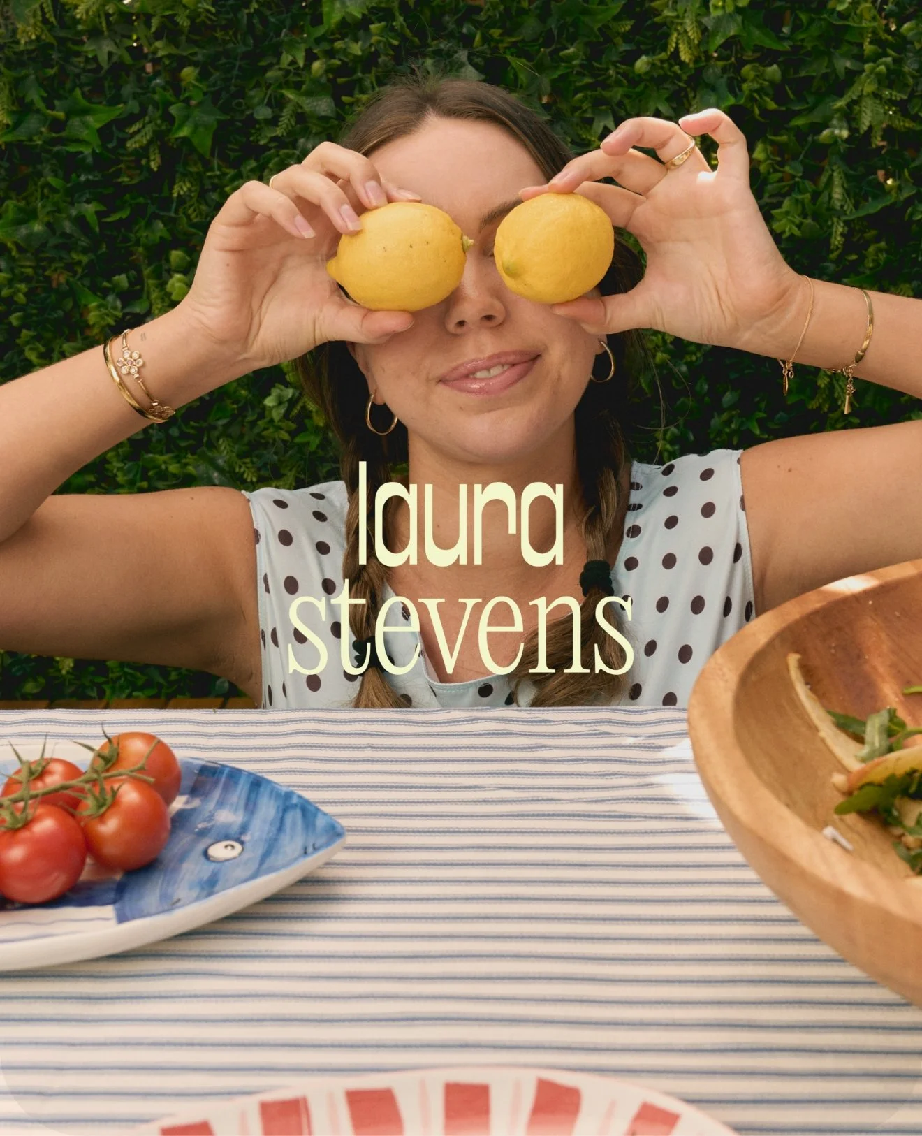 A woman with brown hair and hoop earrings, holding two lemons in front of her eyes, sitting at a table with a striped tablecloth, with tomatoes on a blue and white fish-shaped plate and a wooden bowl with salad in front of her, greenery in the background.