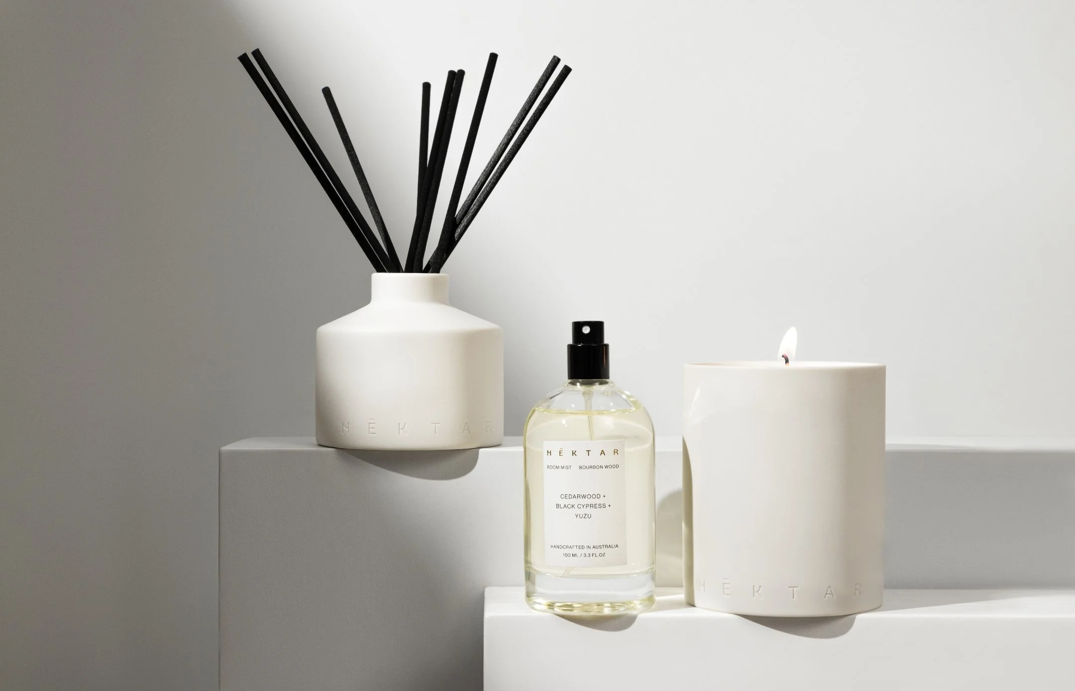 A white vase with black reed diffuser sticks, a clear spray bottle labeled 'NECTAR' with a black cap, and a white candle with a lit wick on a white surface against a light gray background.