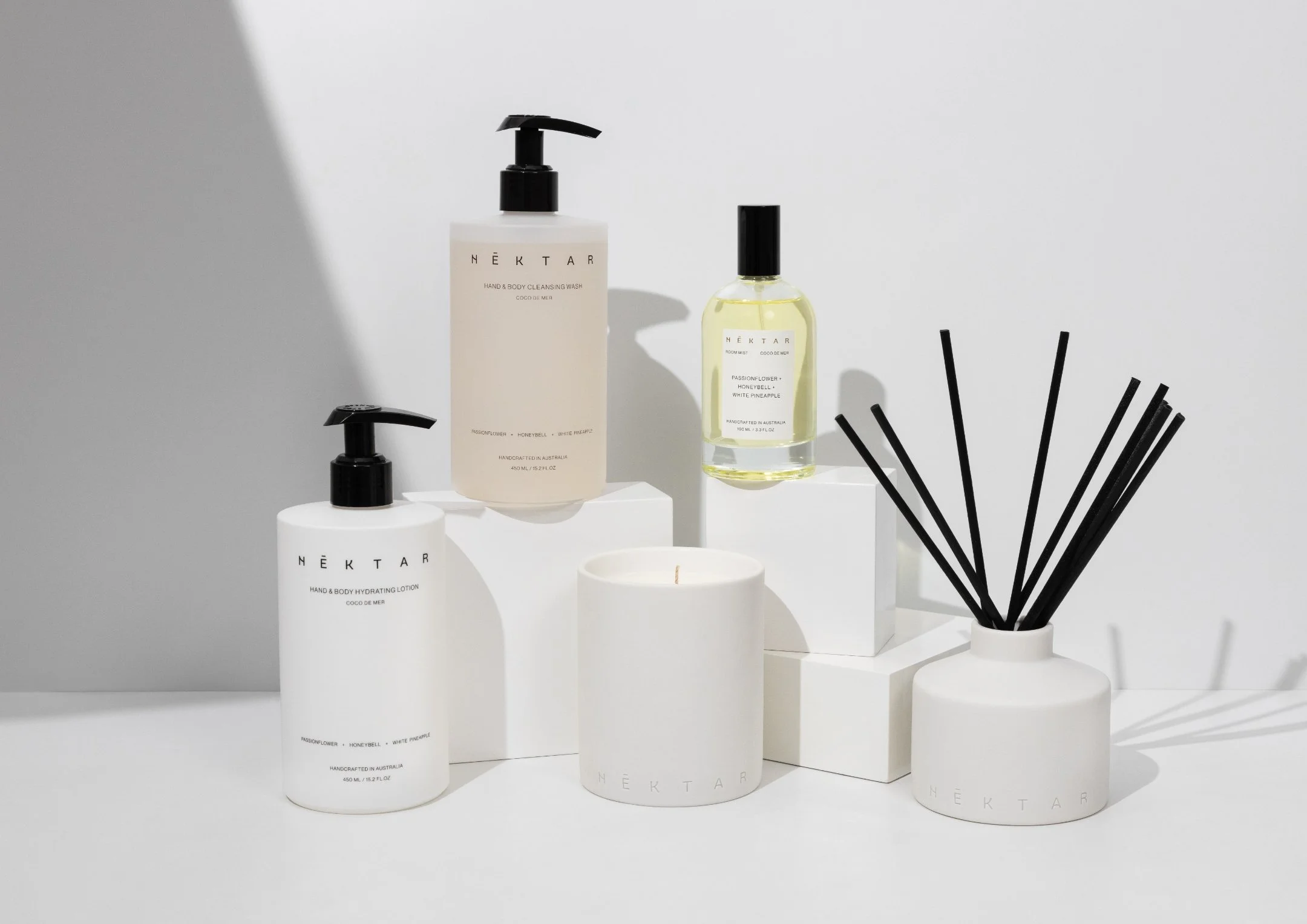 Display of NEKTAR skincare products including pumps and bottles, with a white candle and black diffuser sticks on a white background.