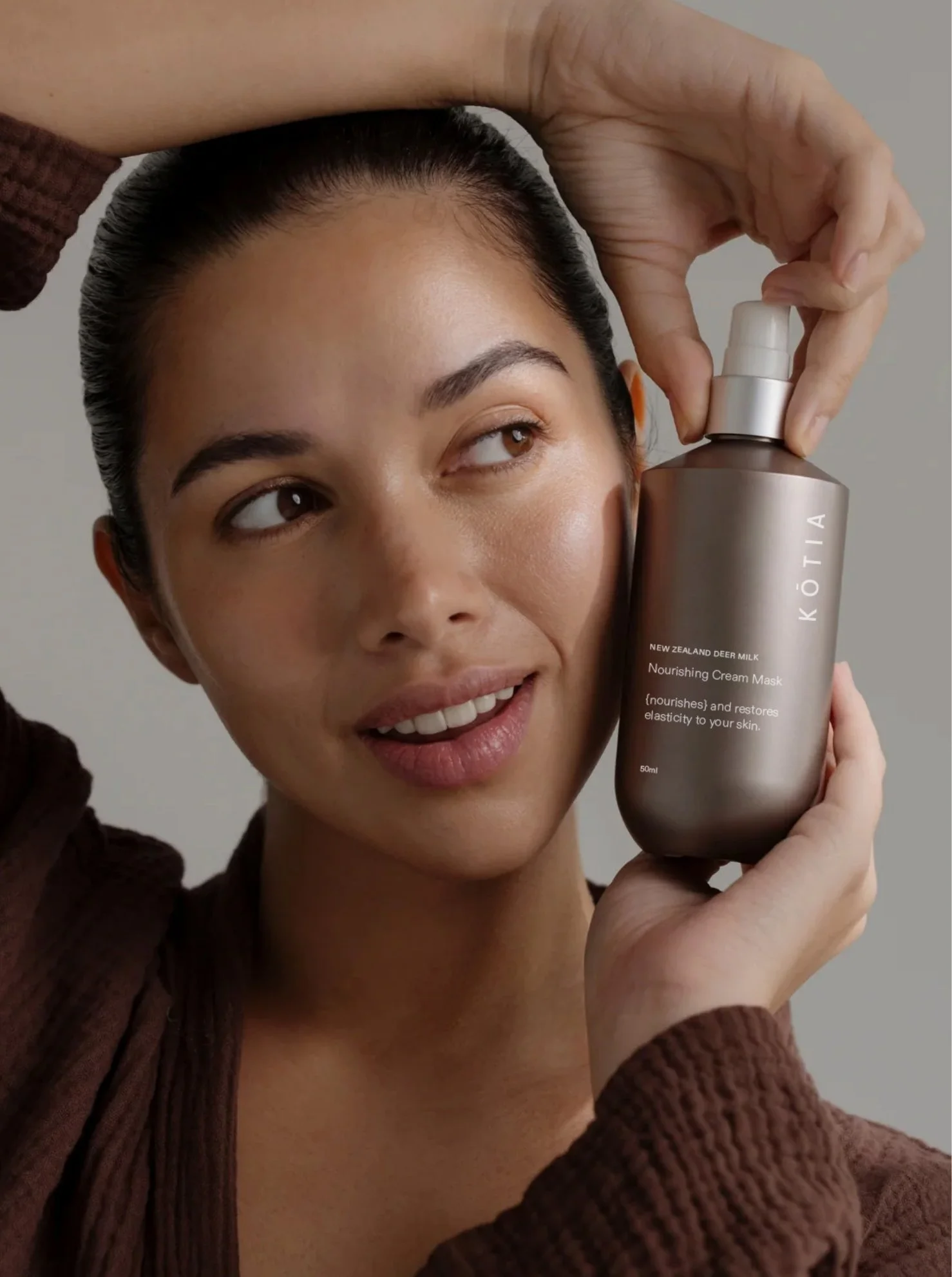 A woman holding a bottle of KŌTIA nourishing cream mask made with New Zealand deer milk, smiling and demonstrating the product.