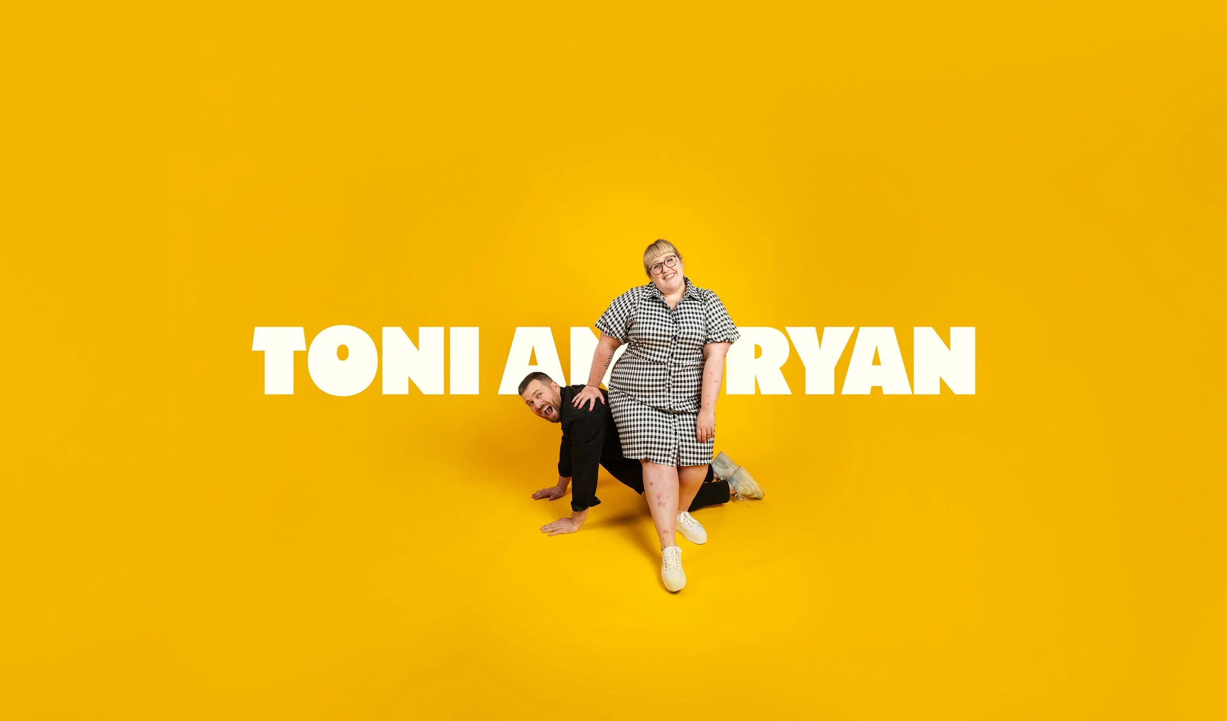 Two people, a man and a woman, are posed in front of a yellow background with the text 'TONI AND RYAN'. The woman, standing, wears glasses and a black-and-white gingham dress, smiling, while the man, kneeling on all fours, is dressed in black and appears to be silly or playful.