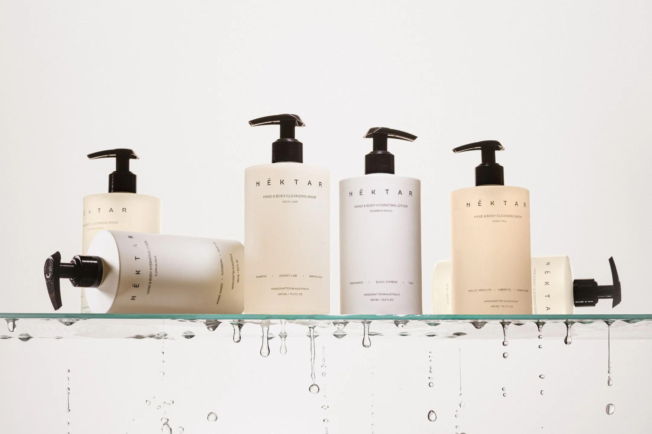 A set of five white and cream skincare bottles with black pump dispensers on a glass shelf, with water droplets hanging below.