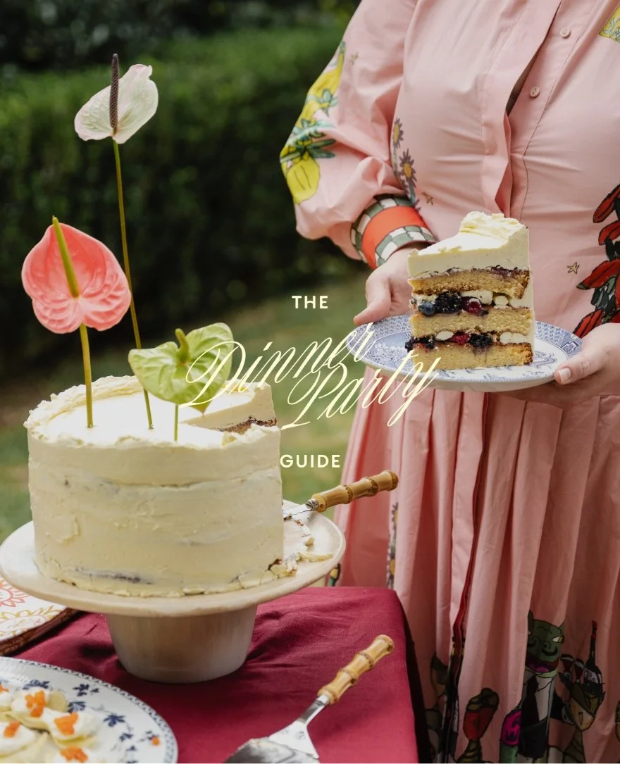 A person in a pink floral dress holding a slice of layered berry cake on a blue plate next to a large white frosted cake decorated with pink and white cake toppers shaped like anthurium flowers, set outdoors on a red tablecloth.