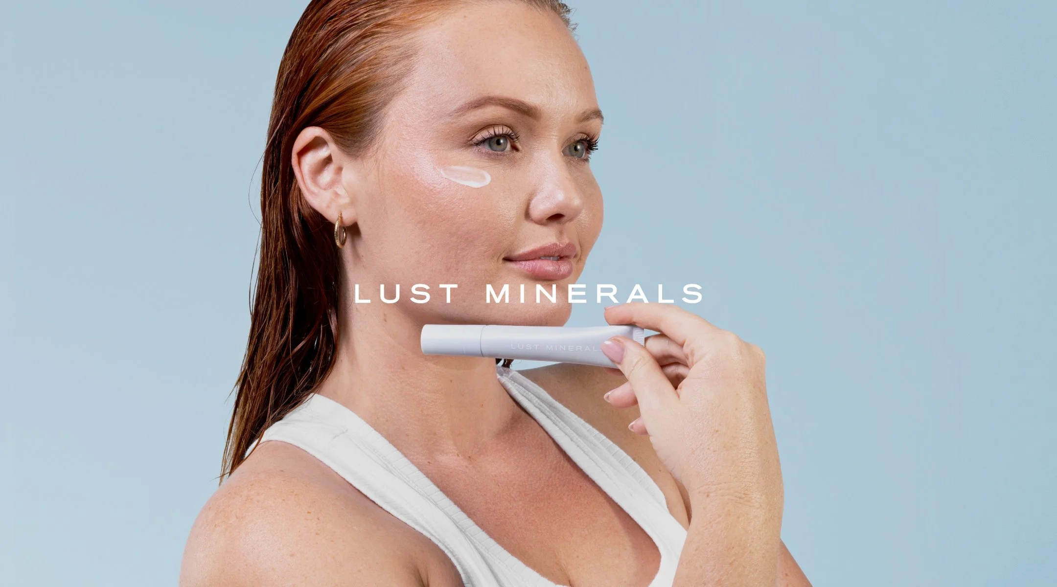 A woman with red hair and earrings is applying a skincare product on her cheek using a white applicator stick. The background is light blue, and the words 'LUST MINERALS' are displayed across the image.