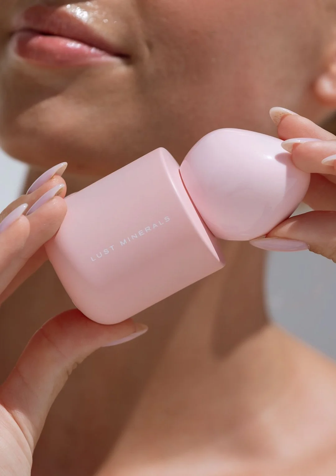 A woman holding a pink Jade roller and a pink box with the words "Lust Minerals" printed on it, near her face.