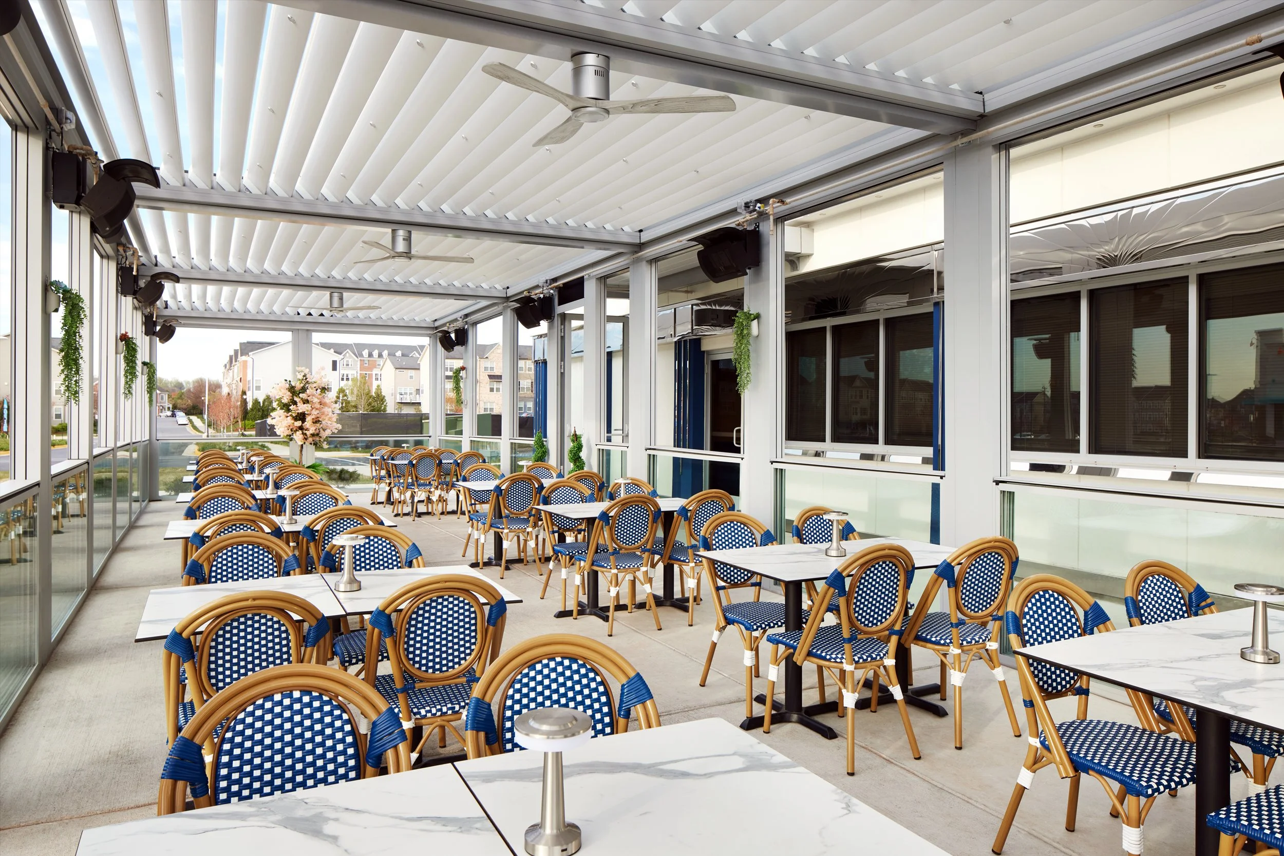 Empty outdoor restaurant patio with blue and white checkered chairs and white marble tables, decorated with potted plants and flowers.