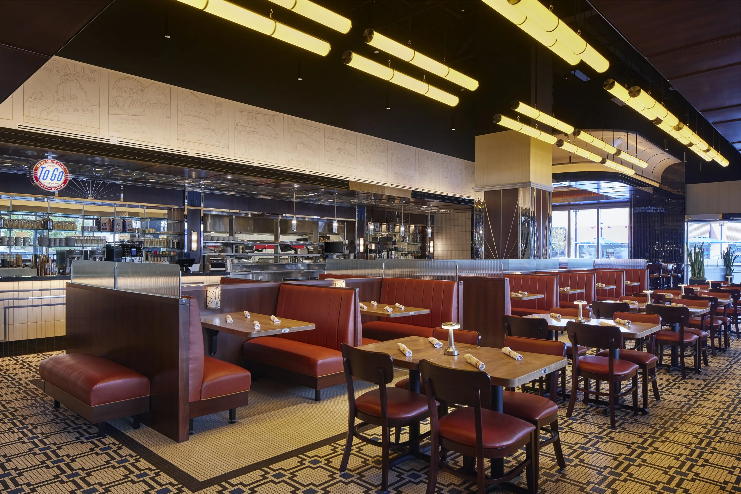 Interior of a modern restaurant with red leather booths, wooden tables set with napkins, and a visible open kitchen area in the background. Overhead lighting is yellow, and large windows allow natural light to enter.