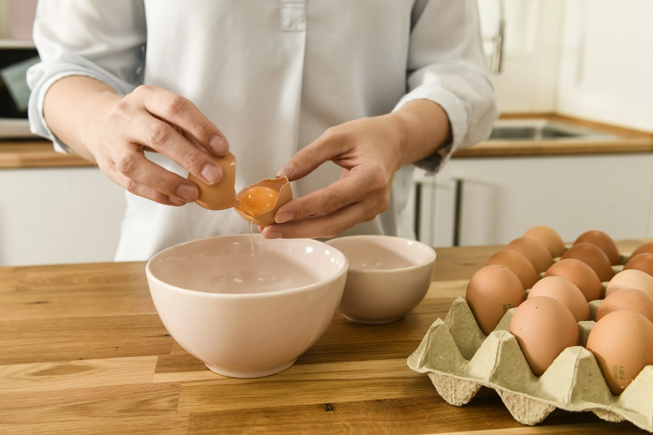 Person cracking a raw egg into a pink bowl on a wooden kitchen countertop, with a carton of eggs nearby.