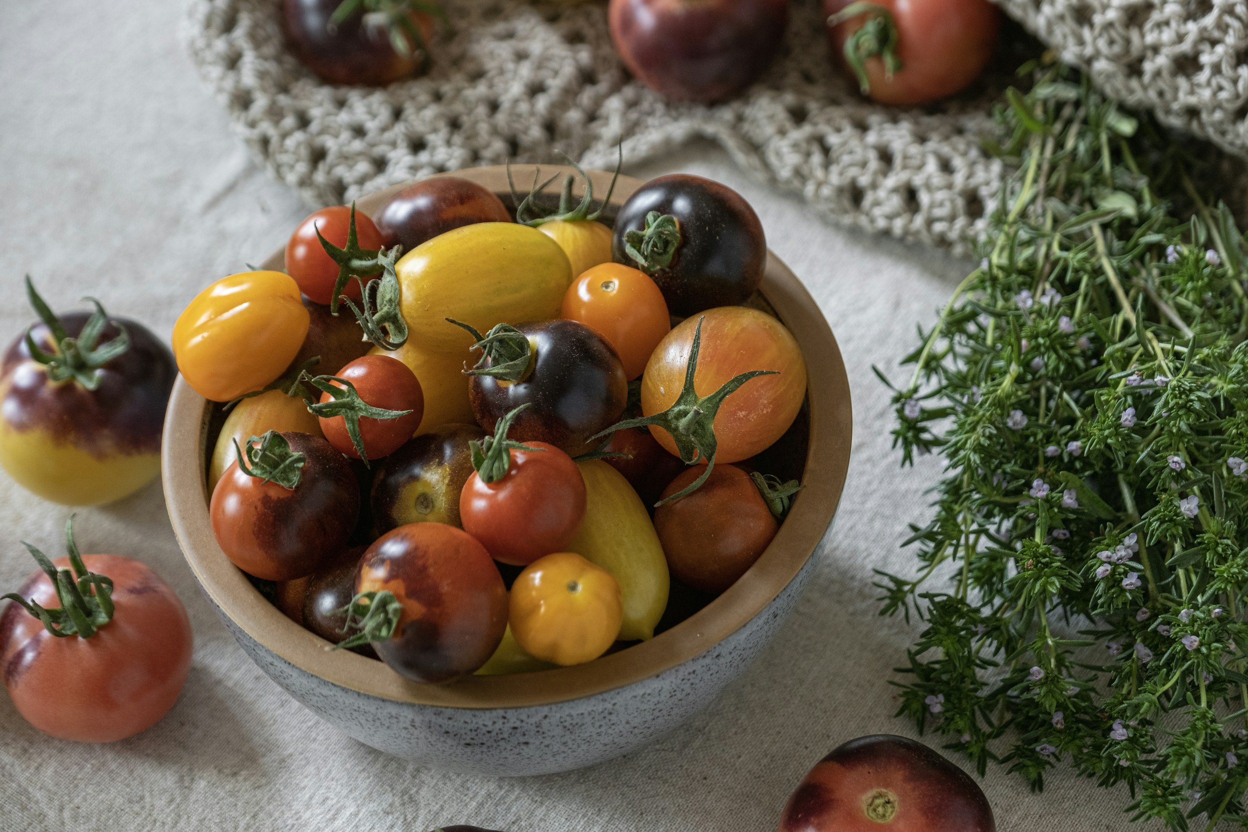 a bowl of heritage tomatoes on a table with green herbs on the side