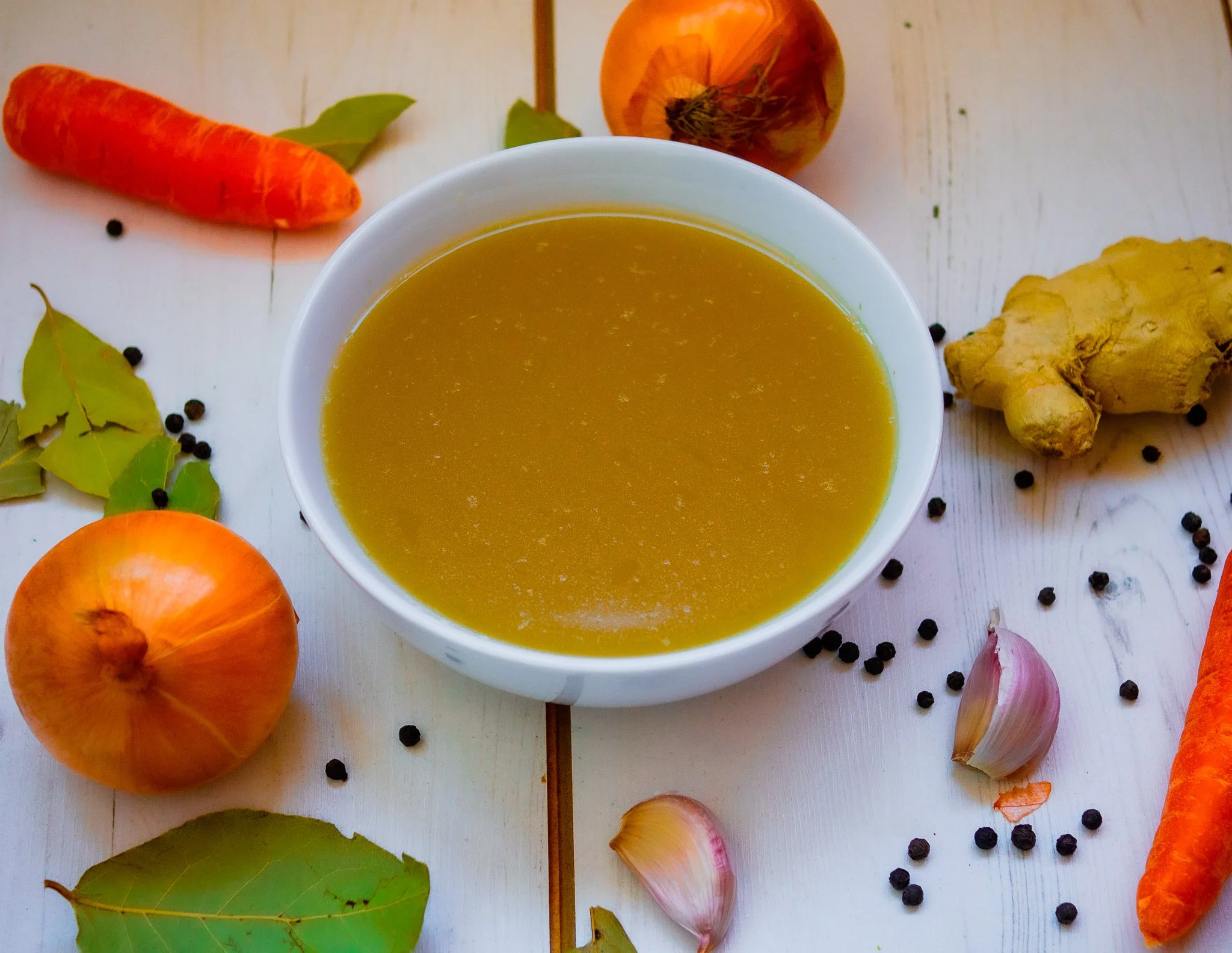 bowl of bone broth in a white bowl surrounded by onions, ginger and carrots