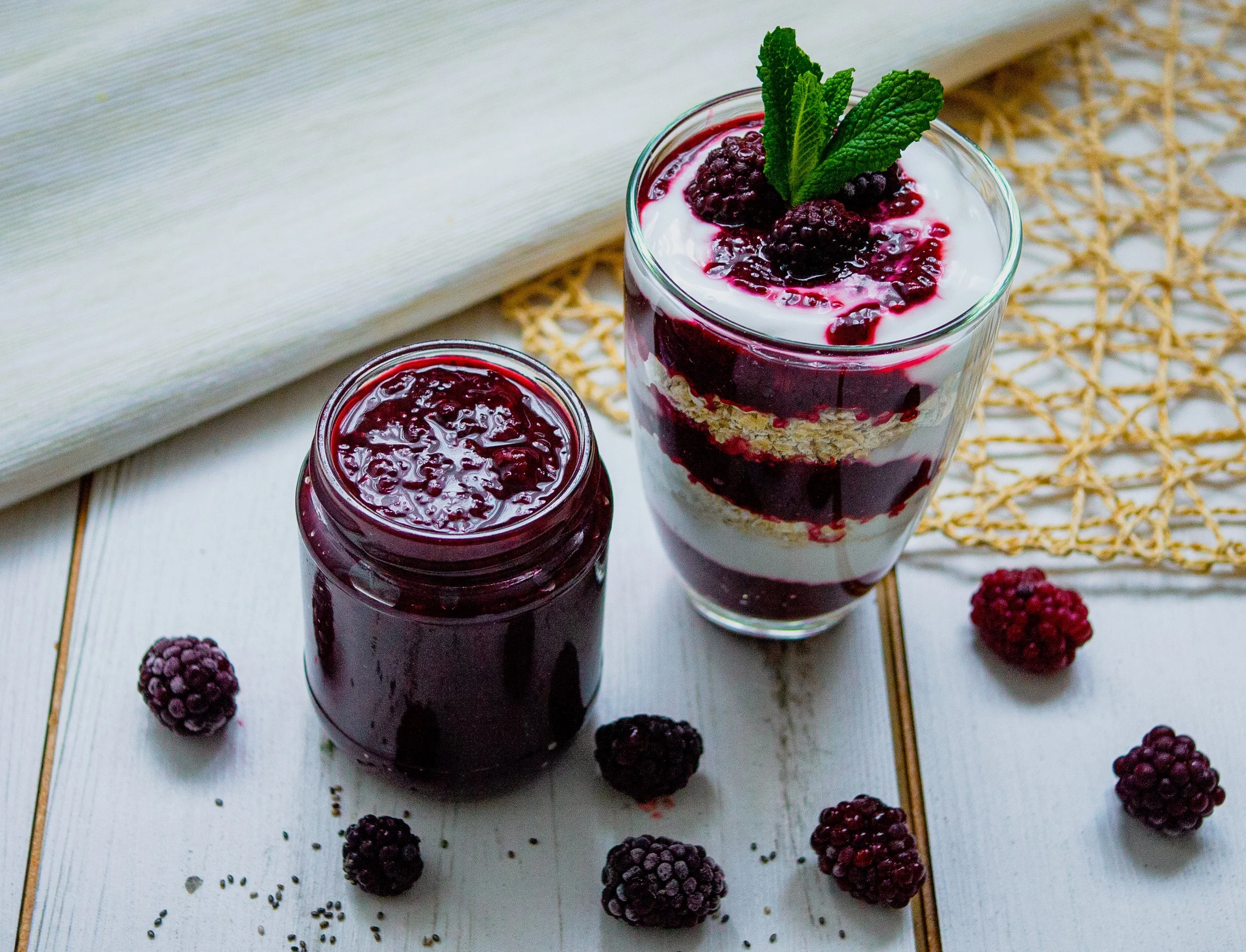 A small jar of blackberry chia jam with a layered yogurt and granola parfait alongside it