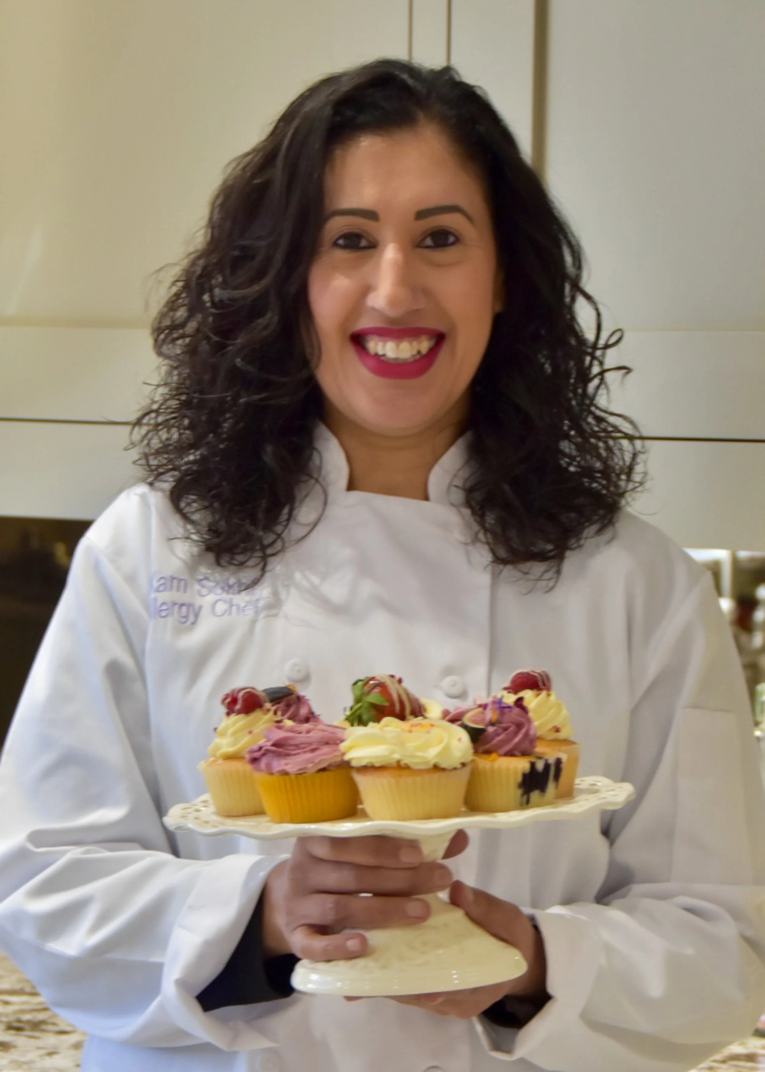 A picture of myself in chef whites holding up a cake stand of co,lourful cupcakes