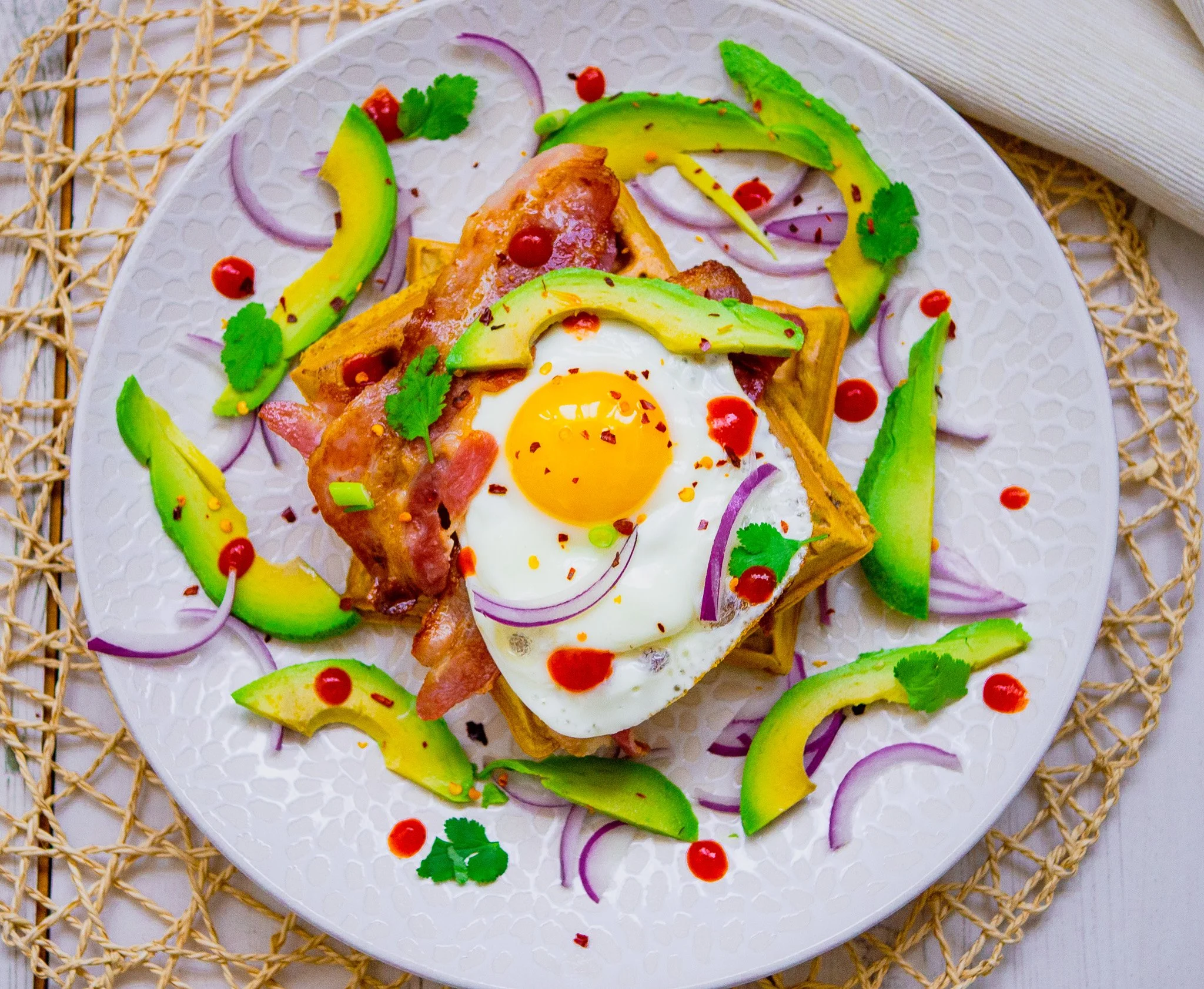 A white plate with savoury waffles topped with avocado, eggs and bacon