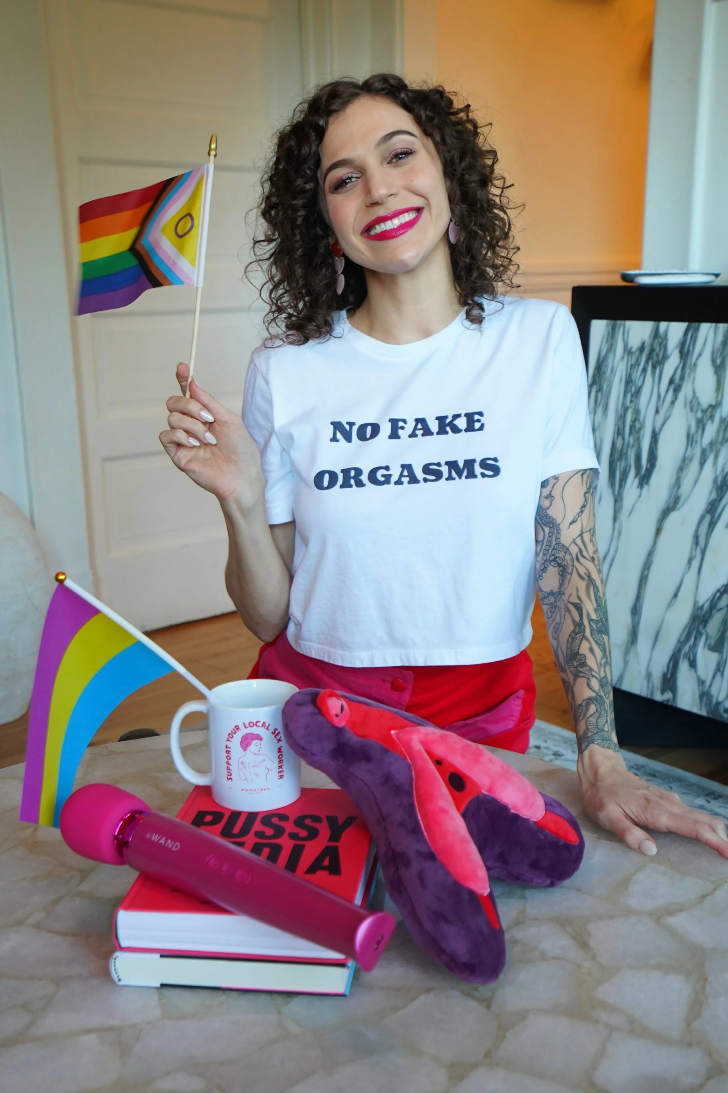 Cady S. Moore, queer sex educator and coach, smiling and holding an intersex-inclusive pride flag while wearing a No Fake Orgasms crop top surrounded by sex education resources